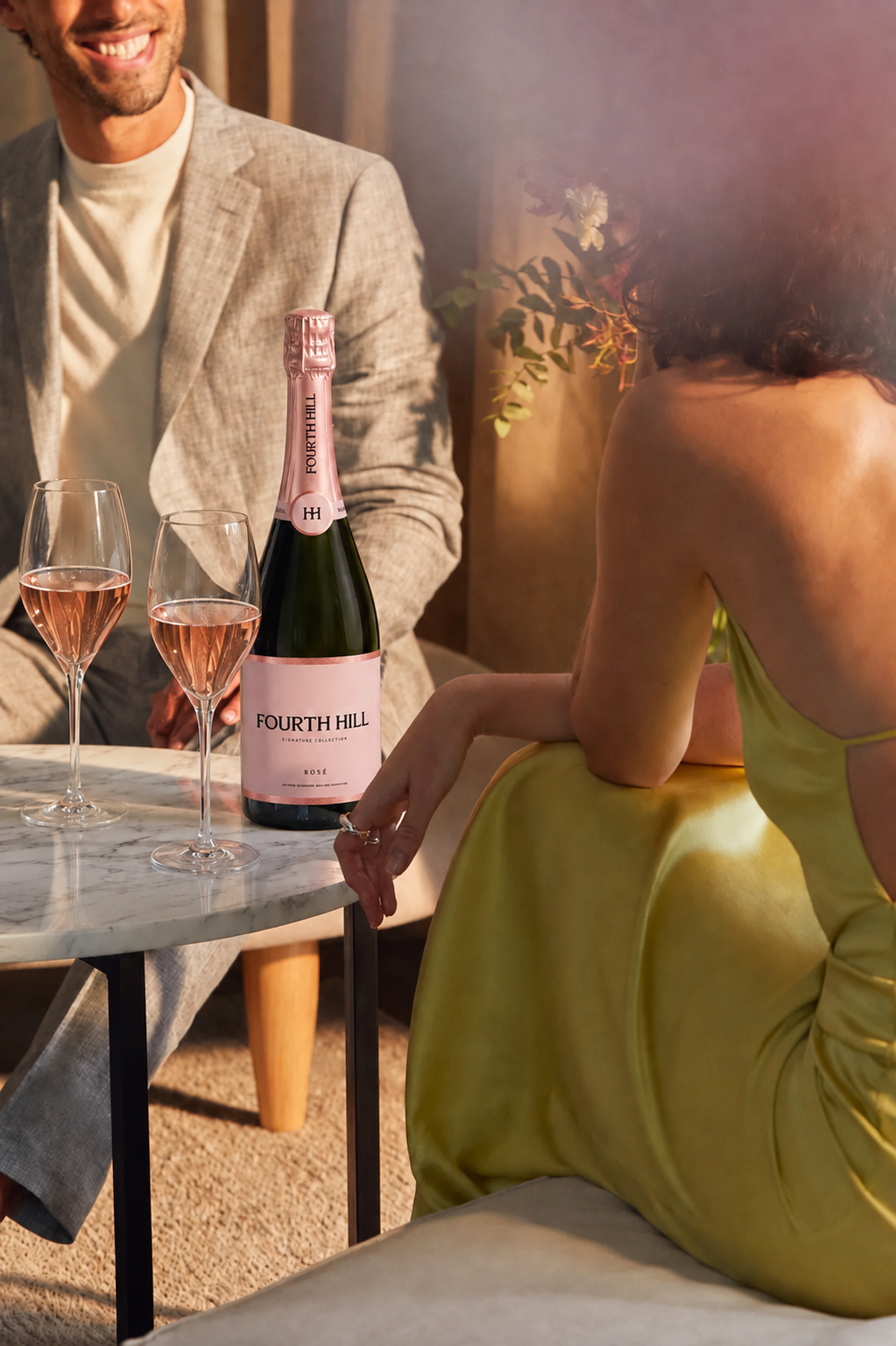 A woman in a yellow satin dress sitting at a marble table with a man in a beige suit. On the table are two filled champagne glasses and a bottle of Fourth Hill Rosé champagne.