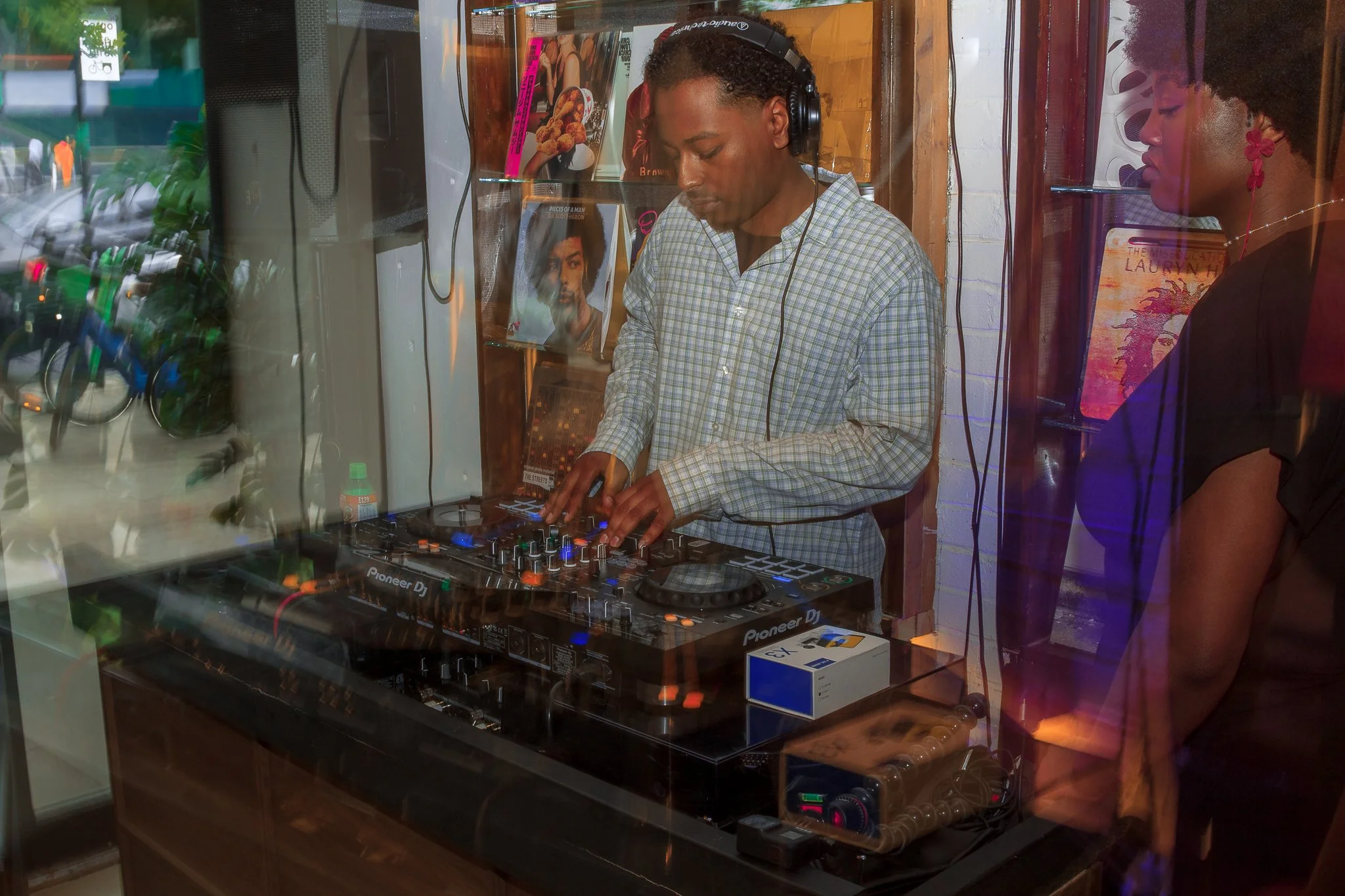 A man DJing with a Pioneer DJ controller, wearing a checkered shirt and headphones, inside a store with books and posters behind him. A woman with curly hair and red earrings watches nearby.
