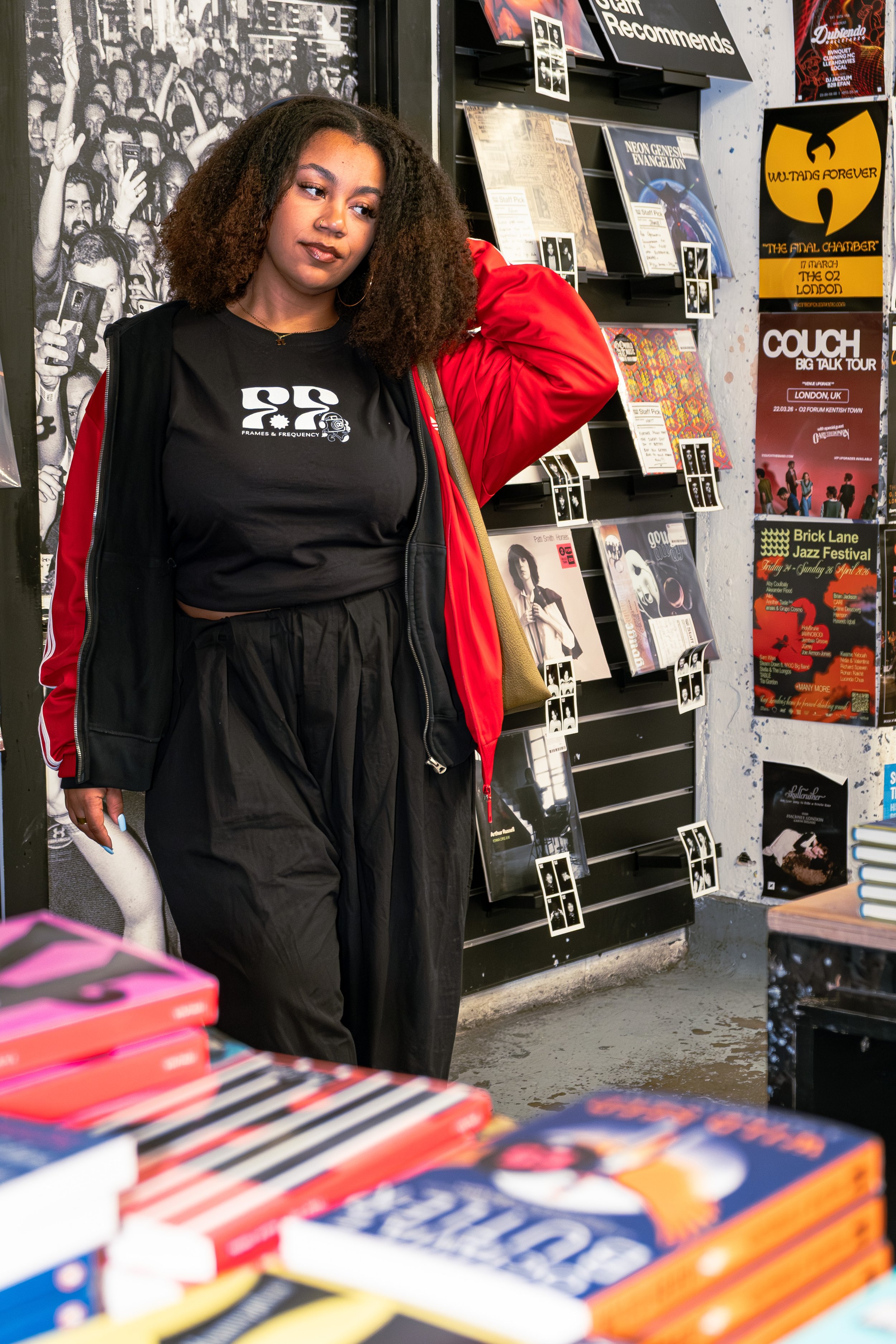 A young woman with curly hair wearing a black t-shirt and a red jacket standing inside a record store, surrounded by shelves filled with vinyl records and posters.