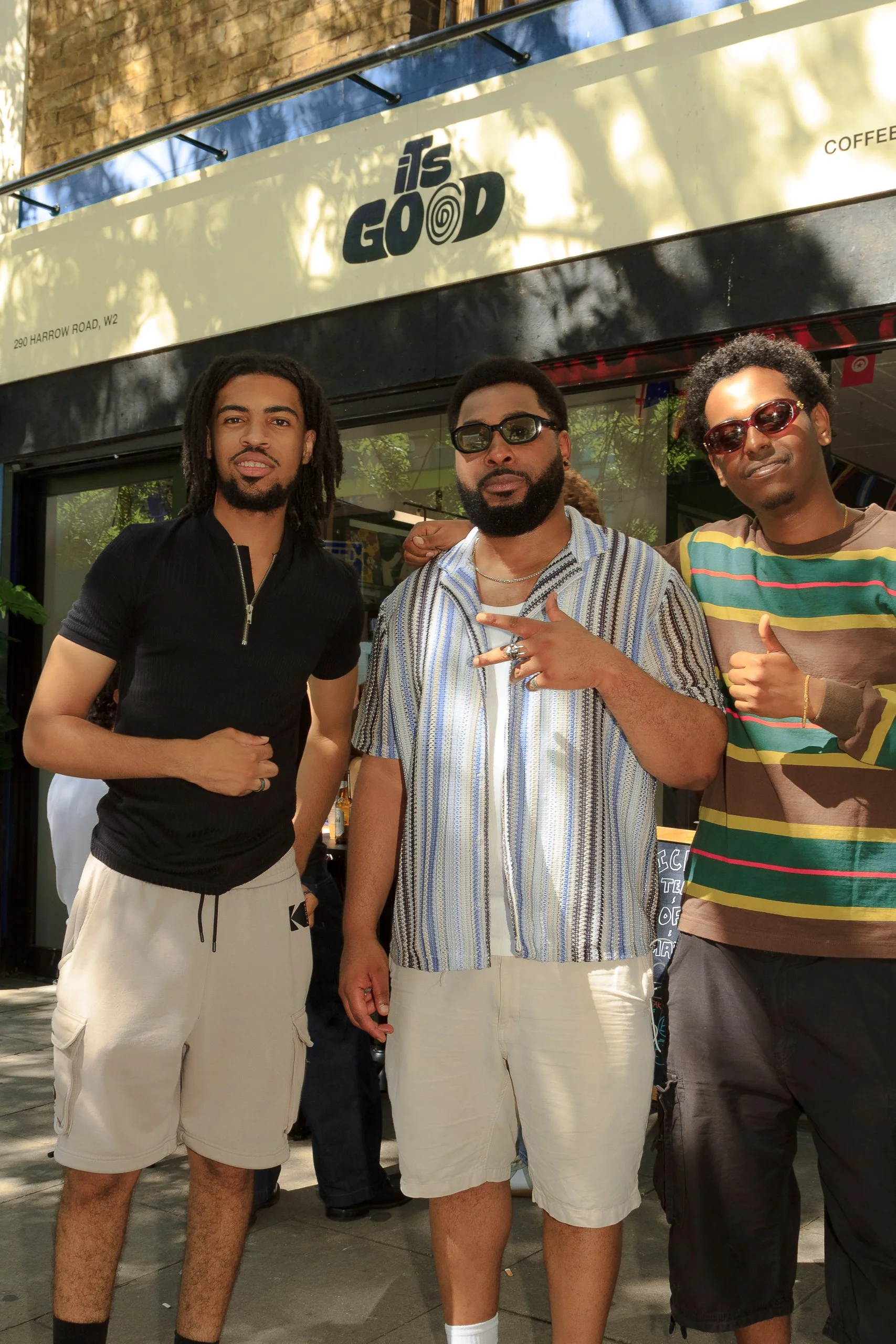 Three men standing outside a cafe or shop, with a sign that reads 'It’s Good' above them. They are dressed in casual summer clothing, and the man on the right is wearing sunglasses.