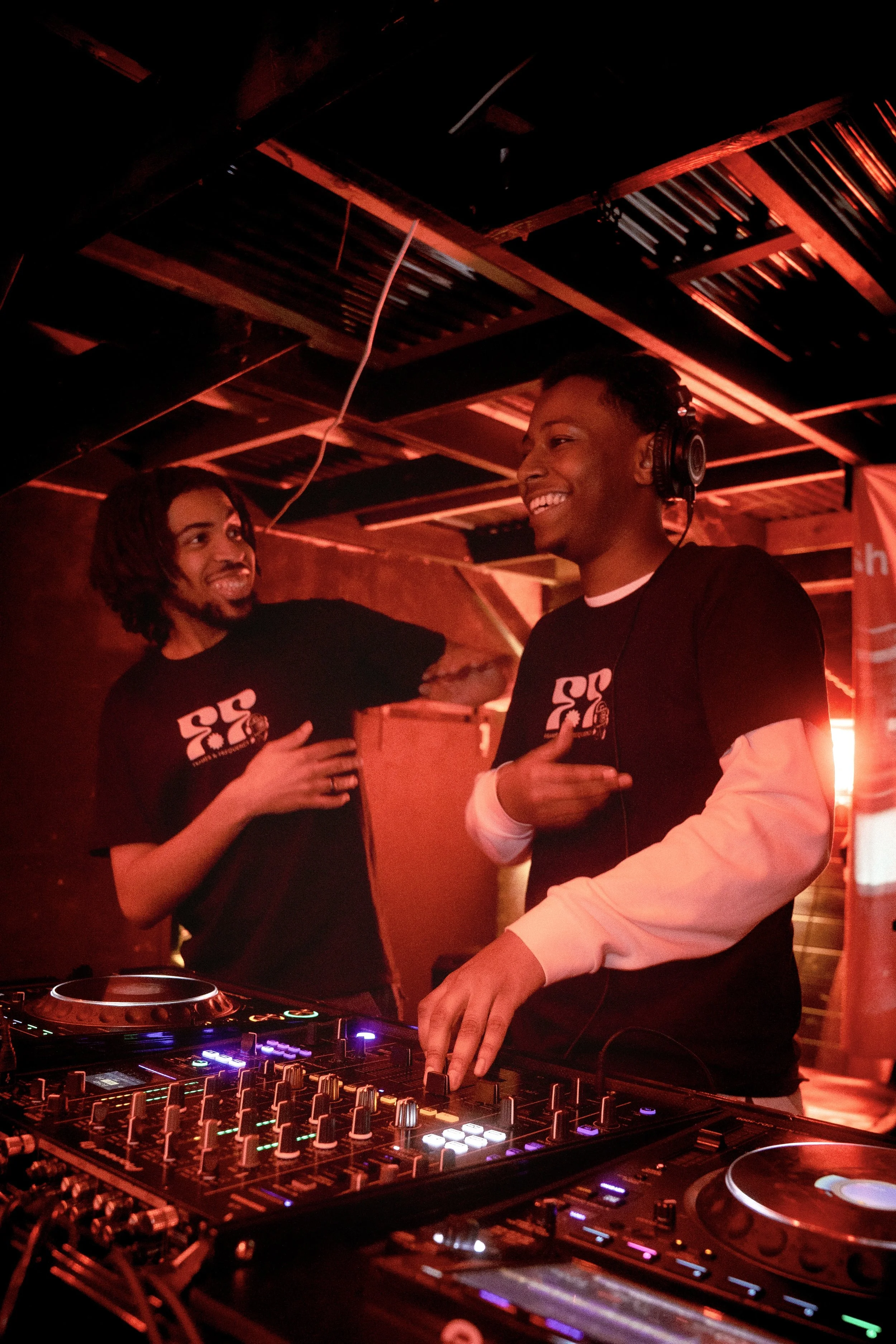 Two men with headphones smiling and talking behind a DJ mixer in a room with red lighting and a wooden ceiling.