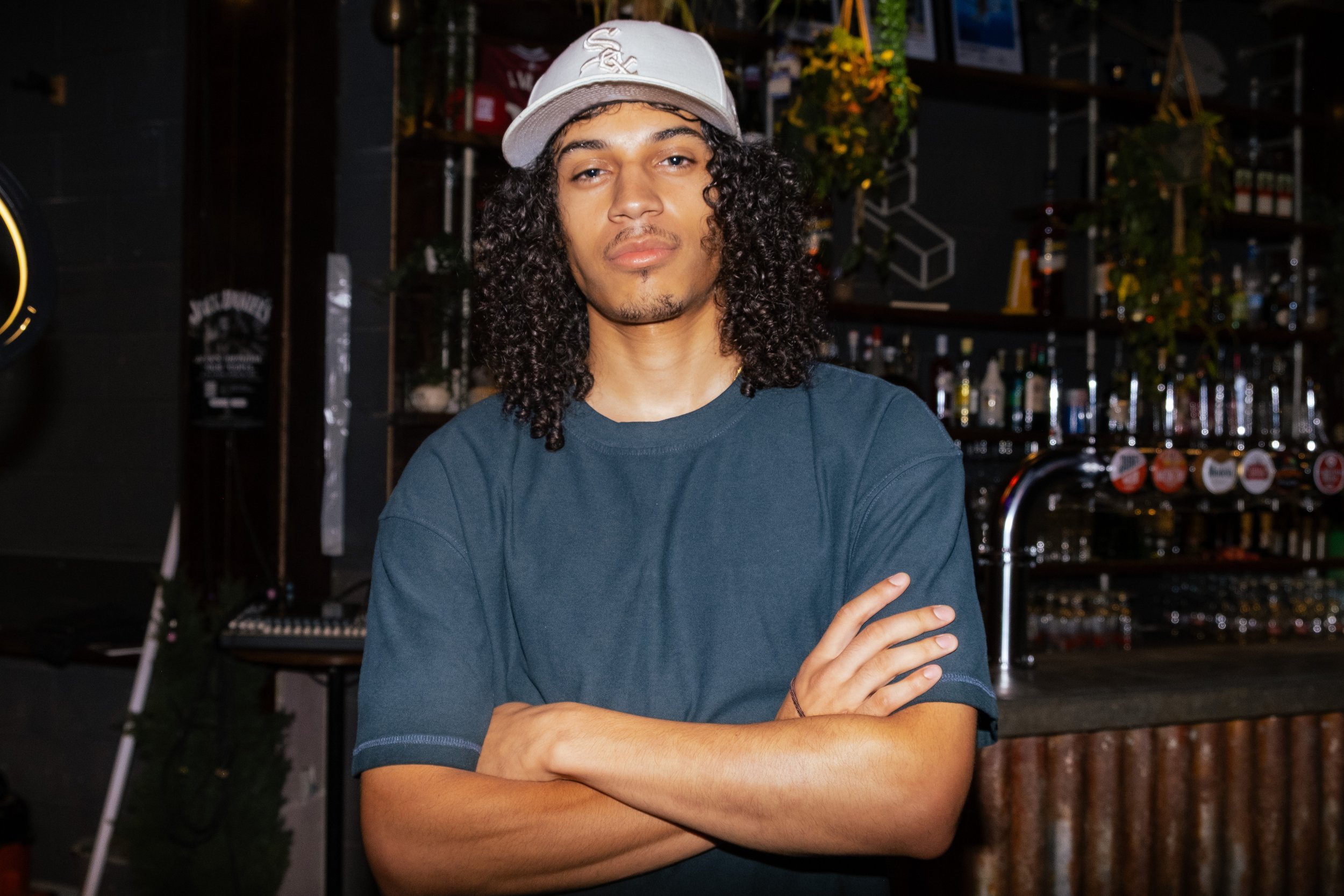 A young man with long curly hair wearing a white baseball cap with a logo and a navy blue T-shirt, standing with crossed arms in front of a bar with bottles and glasses in the background.