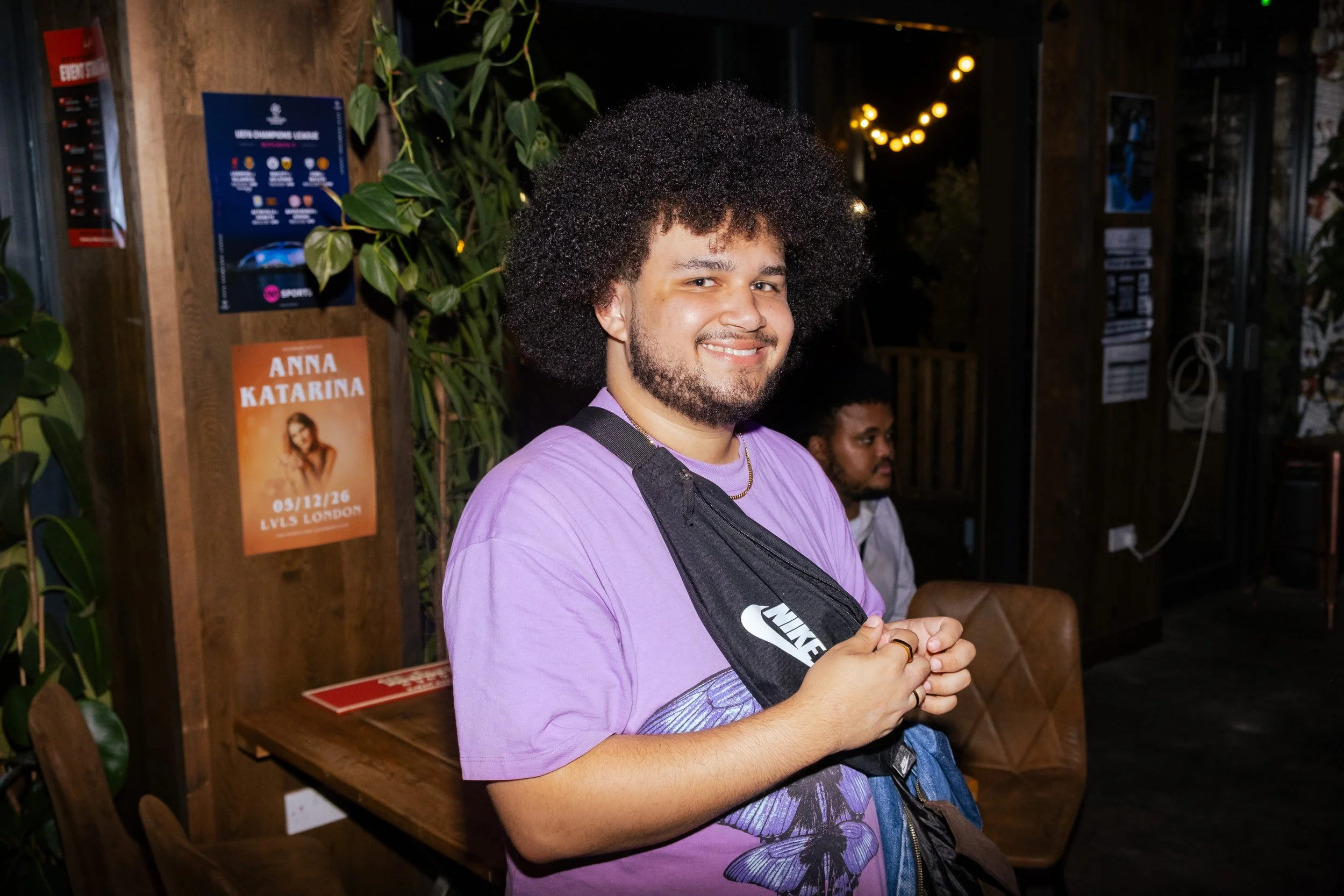A young man with curly hair and a beard is smiling at the camera, wearing a purple shirt and a black Nike crossbody bag. He is standing indoors with a wooden wall and decorative lights in the background. There are posters and another person visible b