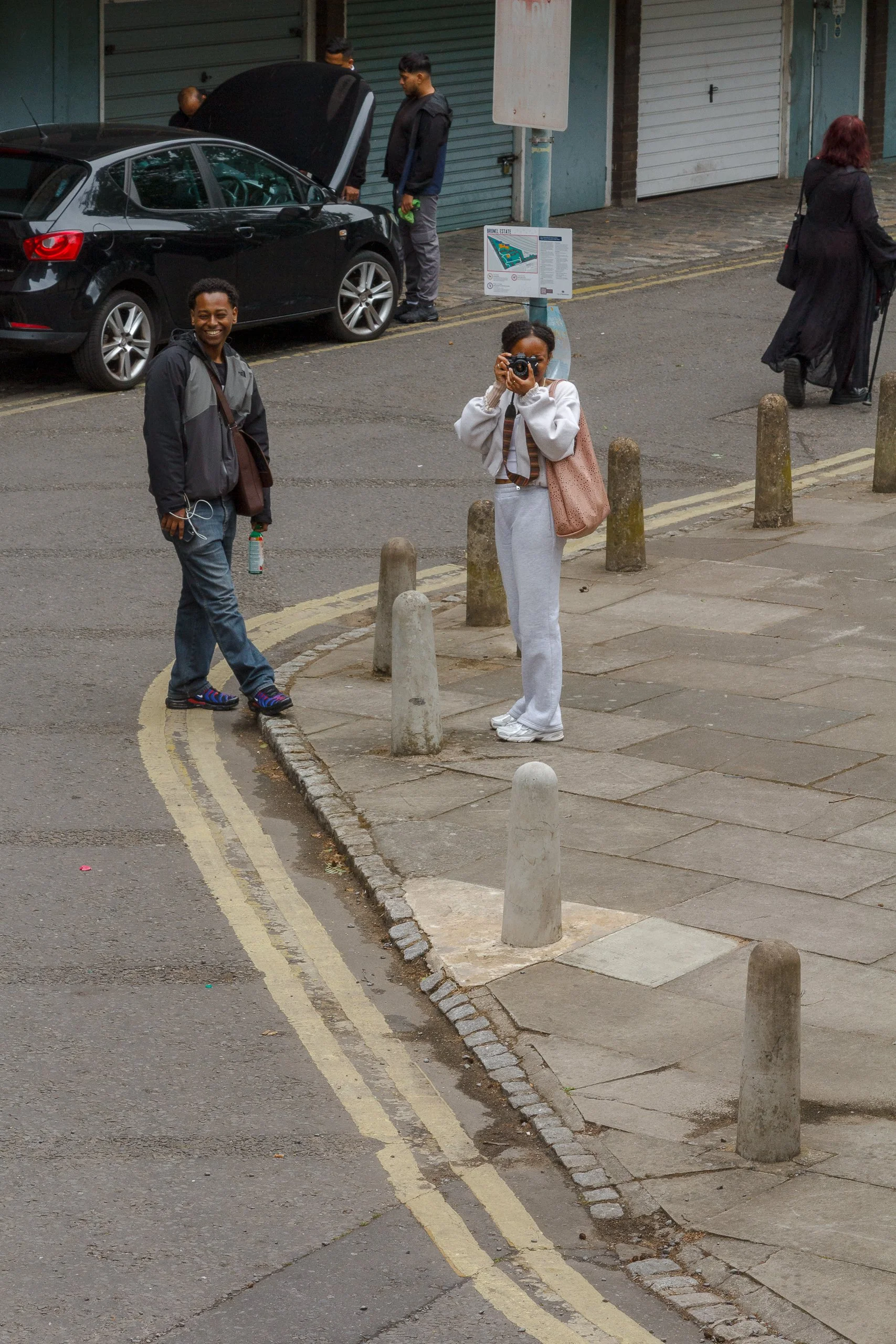 Four people are in an urban setting around a parked black car. One person is smiling and holding a drink, another is looking into the engine of the car, a third is taking a photo, and a fourth is walking away with a suitcase. There are concrete bolla