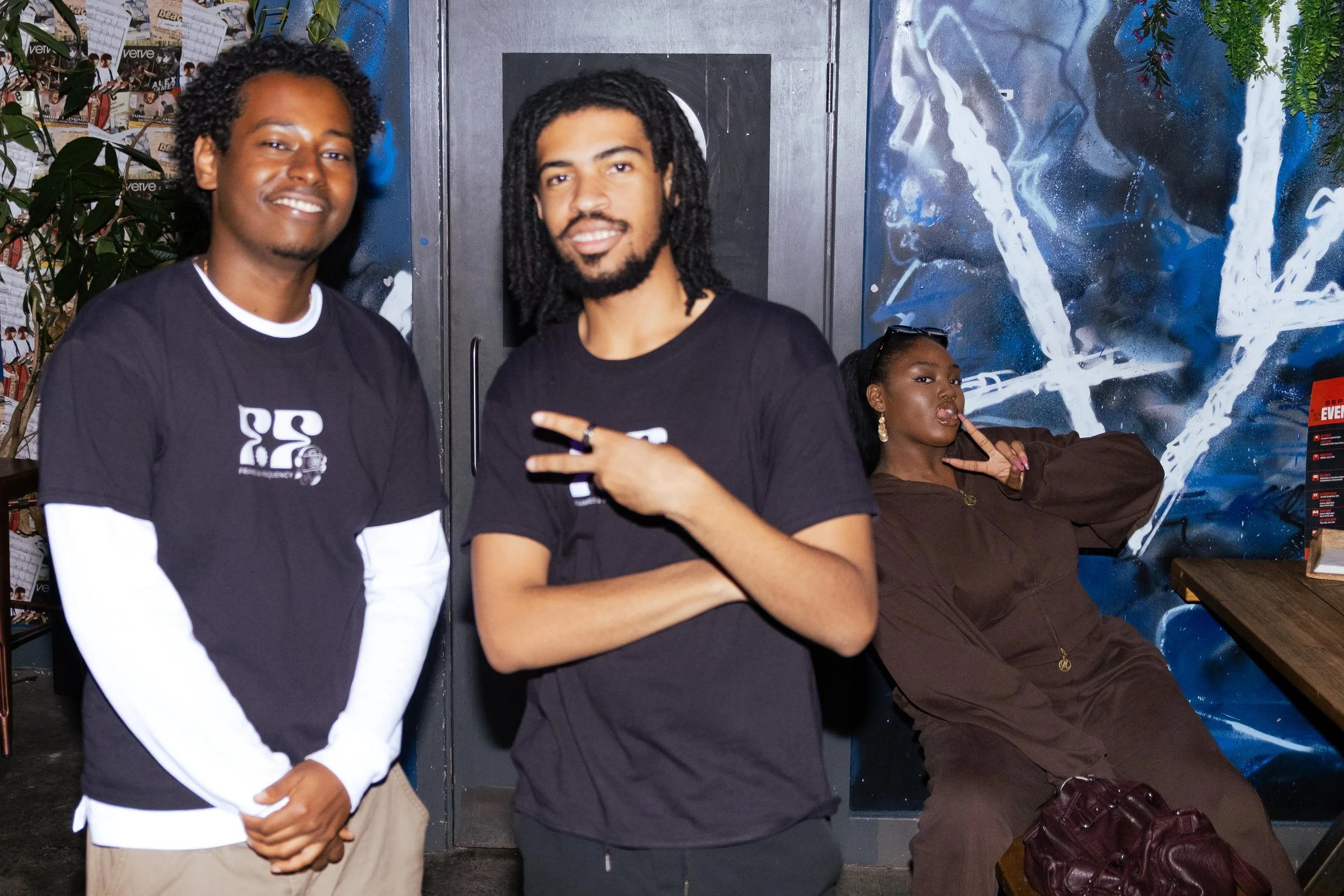 Three people posing indoors in front of colorful abstract wall art. The person on the left has curly hair, wearing a black shirt with white sleeves. The person in the middle has dreadlocks, wearing a black shirt, making a peace sign. The person on th