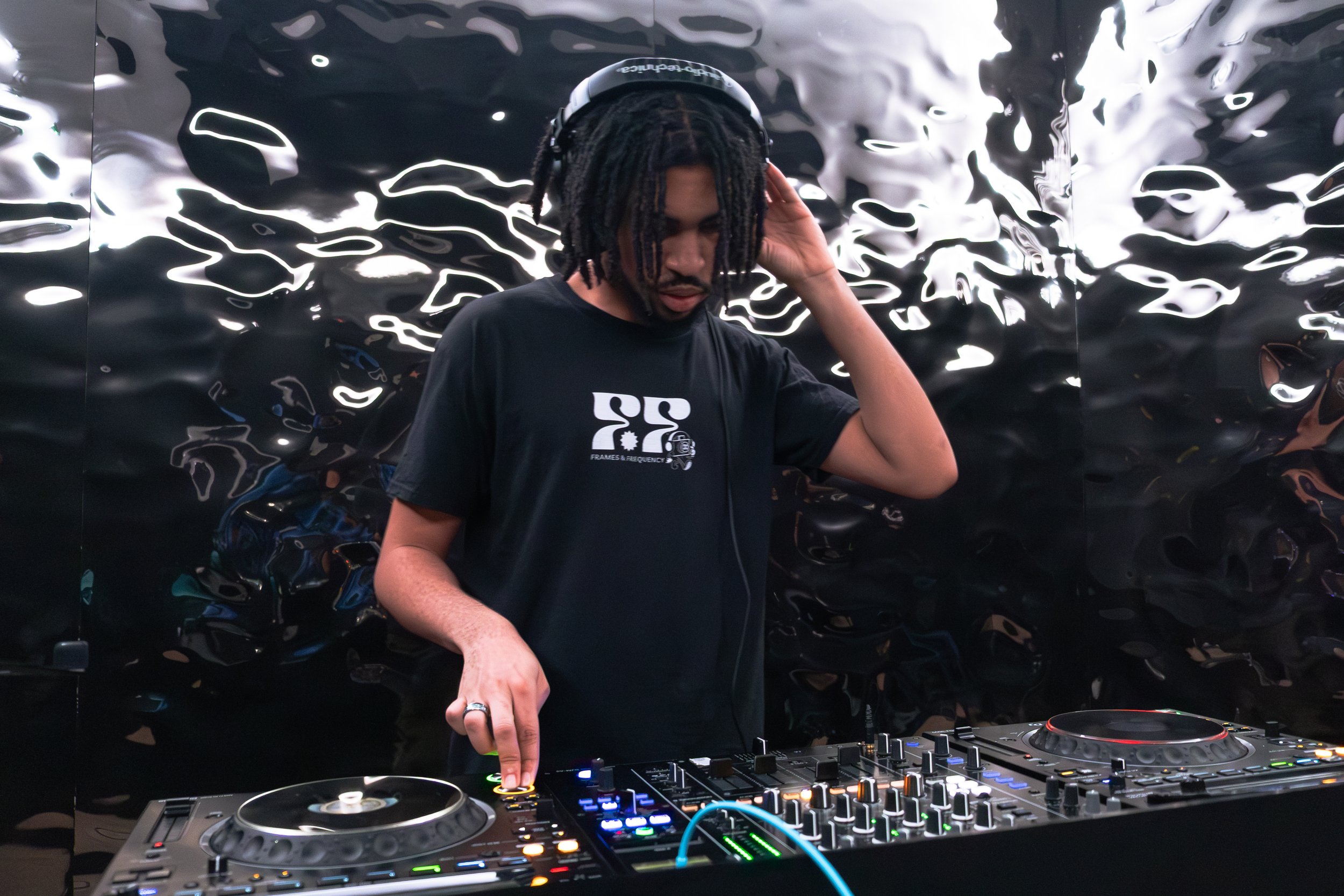 A male DJ with dreadlocks wearing headphones, adjusting equipment at a music setup with a reflective, wavy black background.