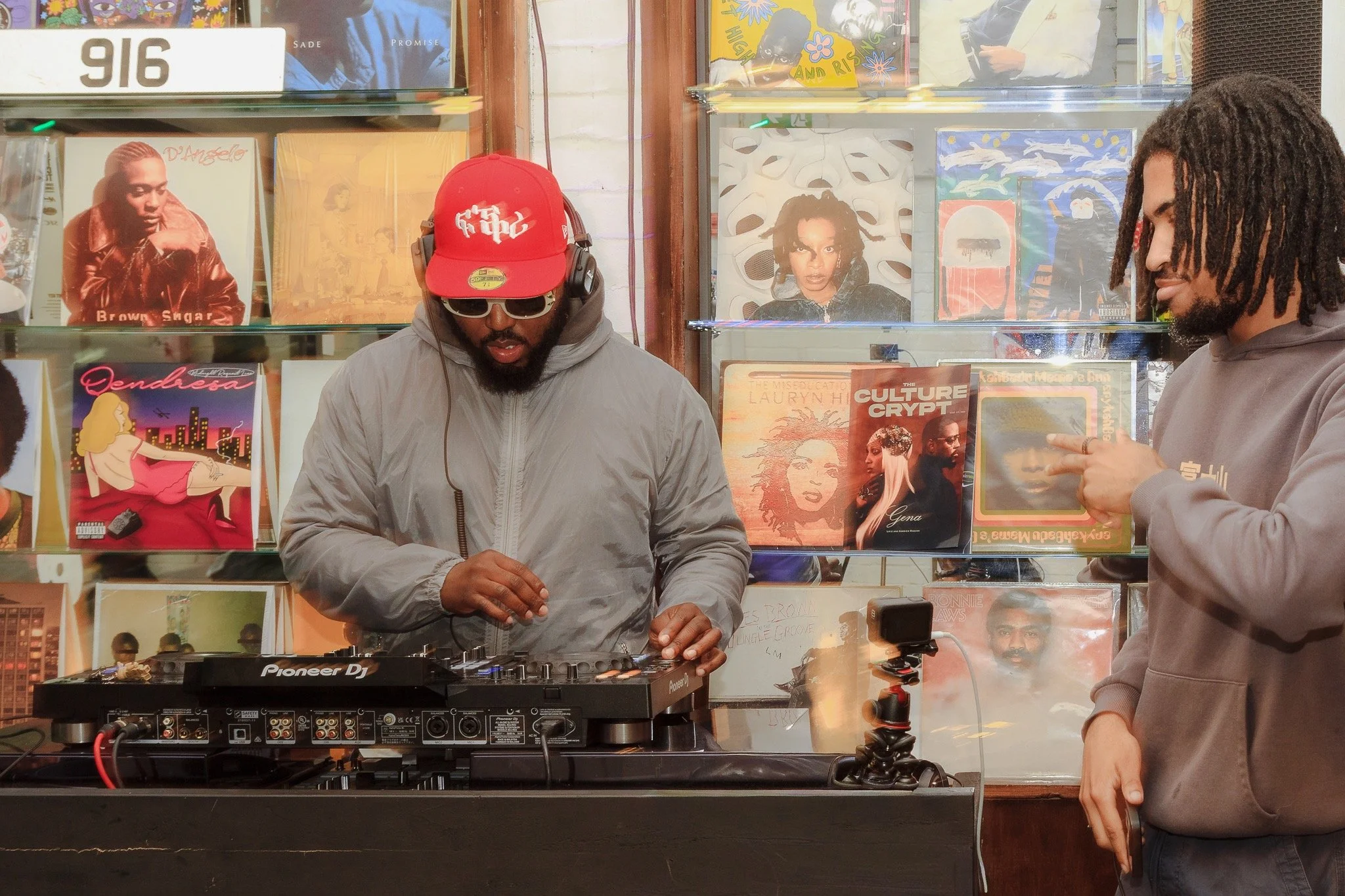 Two men are in a record store, with one using a DJ mixer and wearing headphones, sunglasses, a red cap, and a gray jacket. The other man is on the right side, with dreadlocks, a gray hoodie, and is gesturing with his hand. Behind them are shelves wit