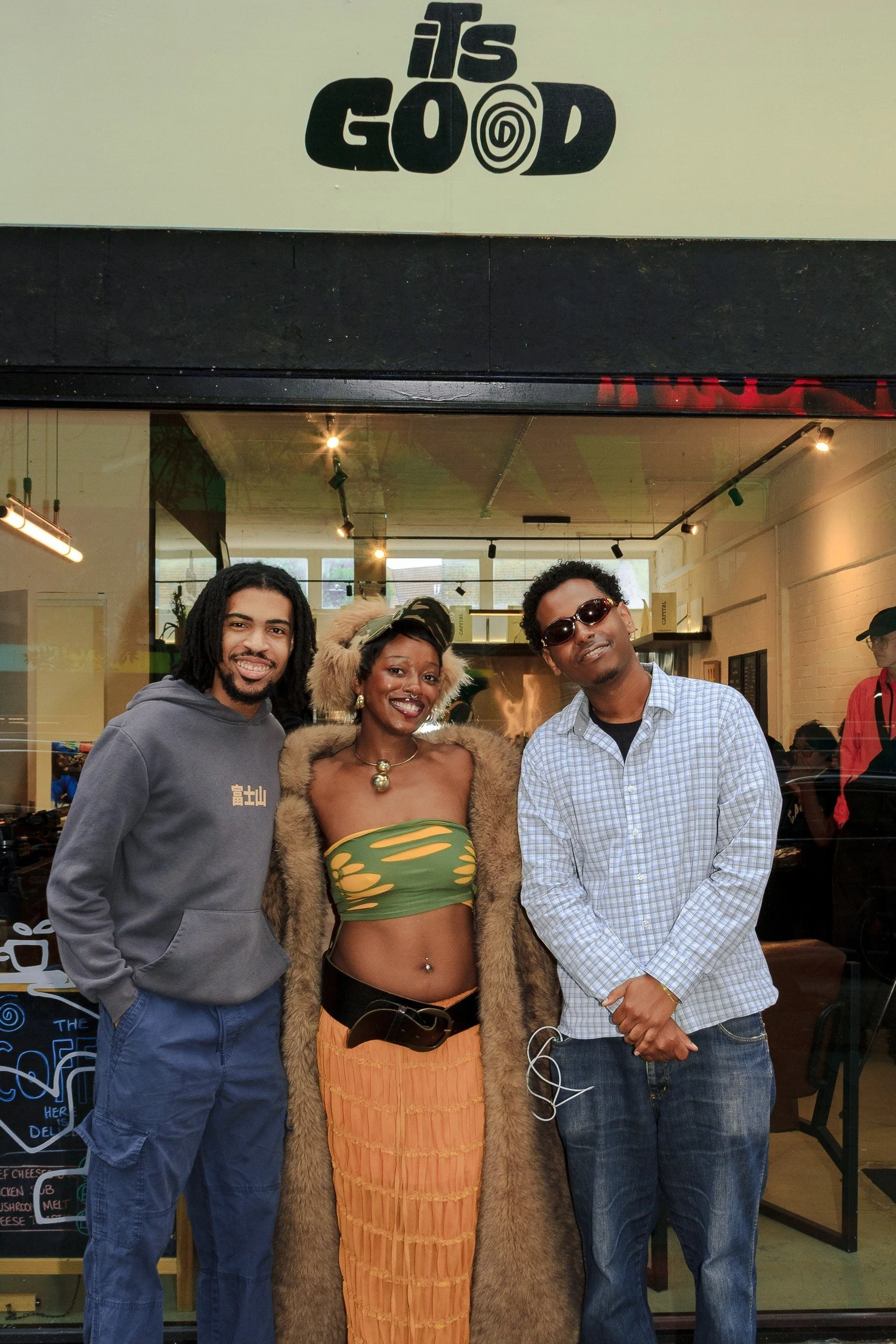 Three people standing together inside a cafe, smiling at the camera. The person in the middle is a woman wearing a fur coat and a green and yellow outfit underneath, with a large necklace. The two men on either side are dressed casually, with one wea