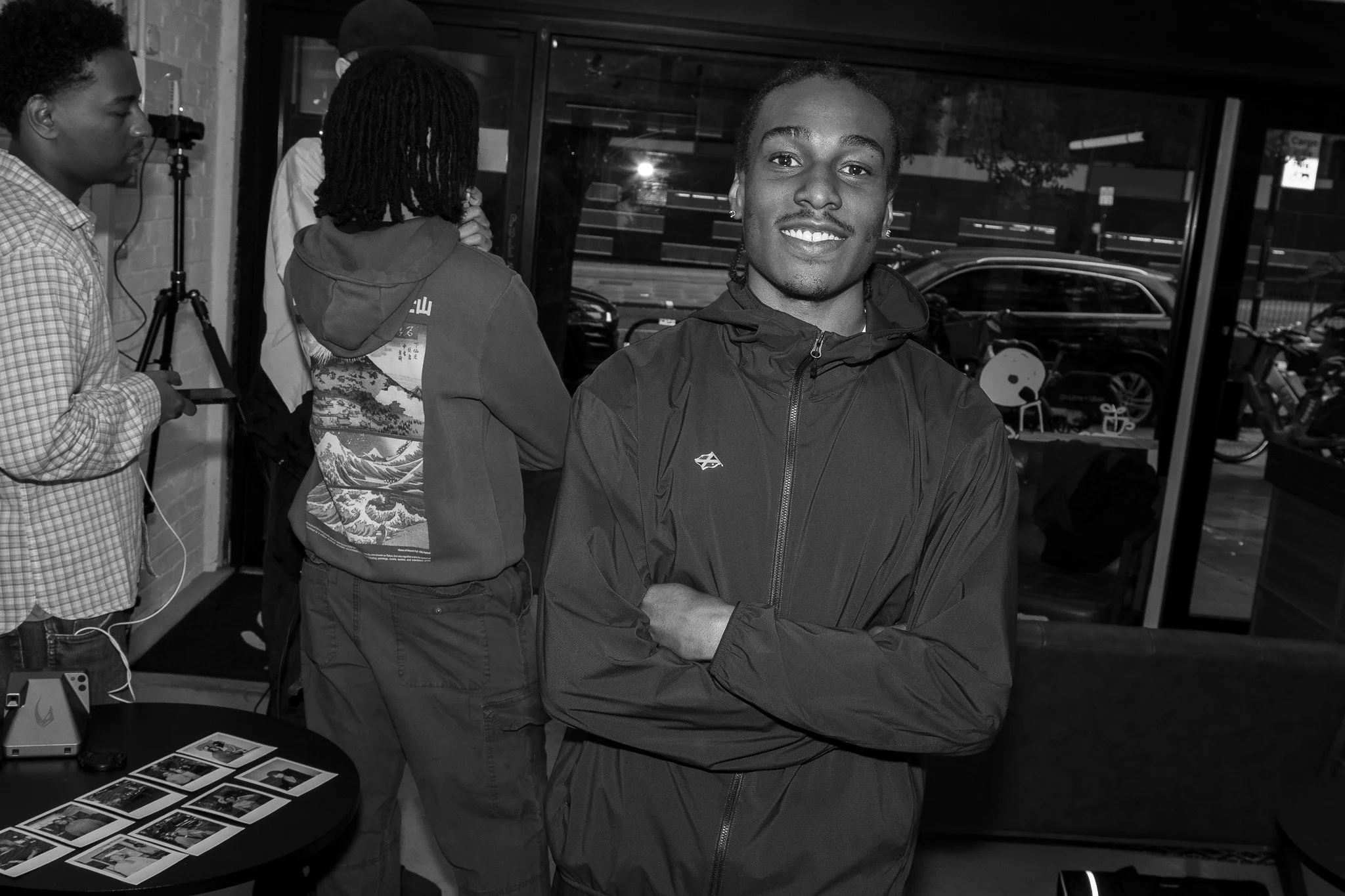 A young man with short curly hair, earrings, and a smile, wearing a dark jacket with a logo, standing with arms crossed inside a store or cafe. In the background, there are three other people, one with dreadlocks and a hoodie, another with short hair
