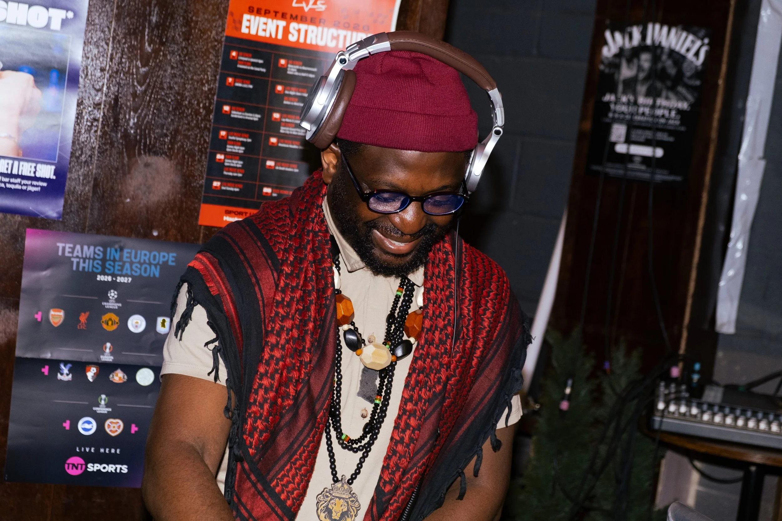 A man wearing glasses, a red beanie, and layered necklaces, standing in front of posters and event information at an indoor venue.