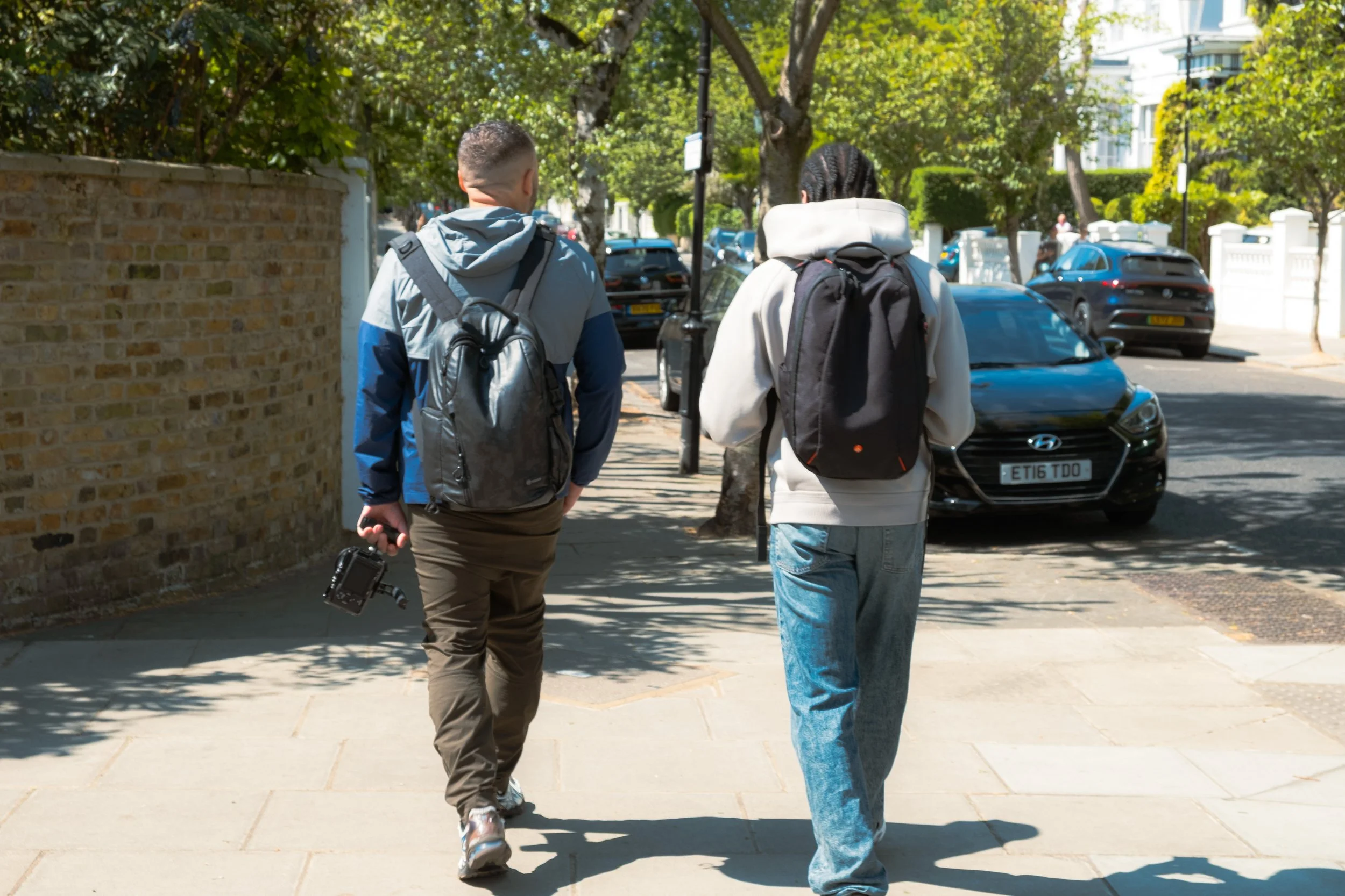 Two people with backpacks walking on a city sidewalk, seen from behind, with parked cars and trees lining the street.