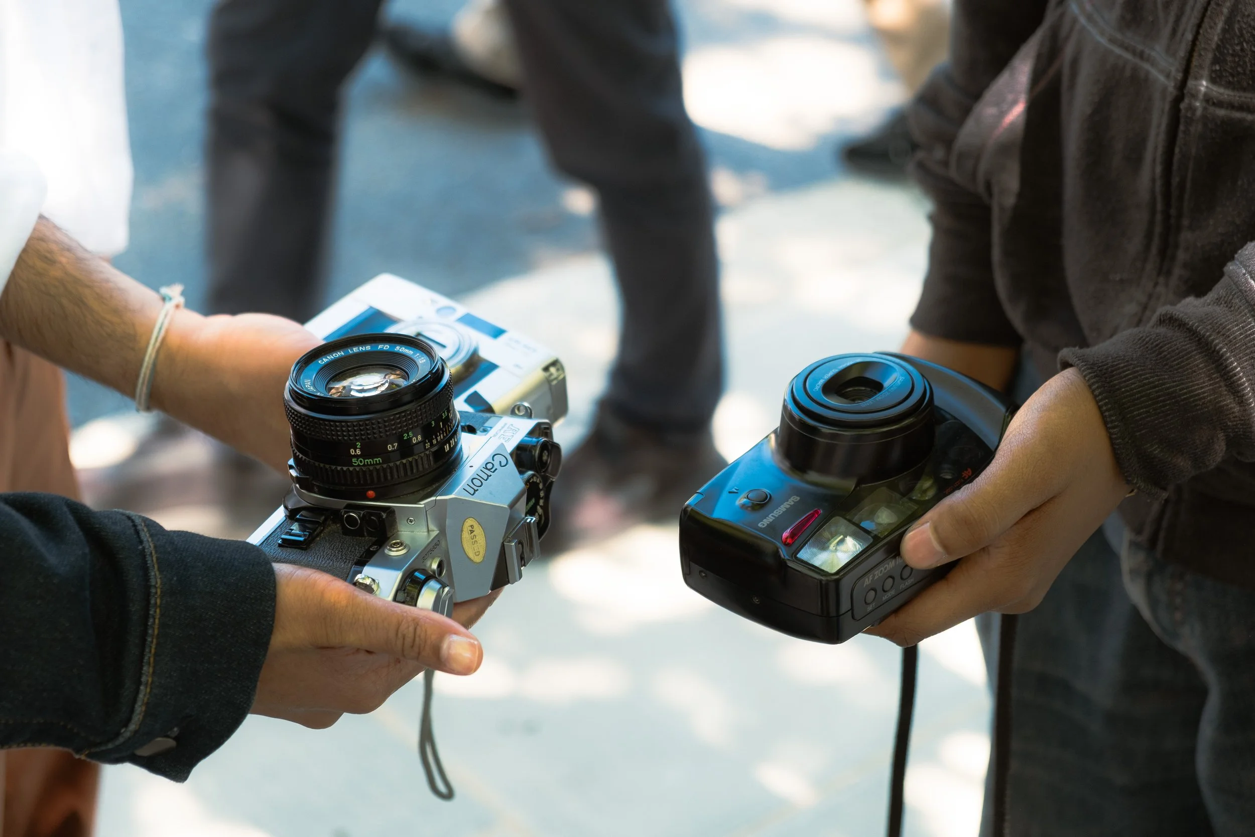 Close-up of two people exchanging a camera and a camera flash, with a blurred background showing legs and shoes.