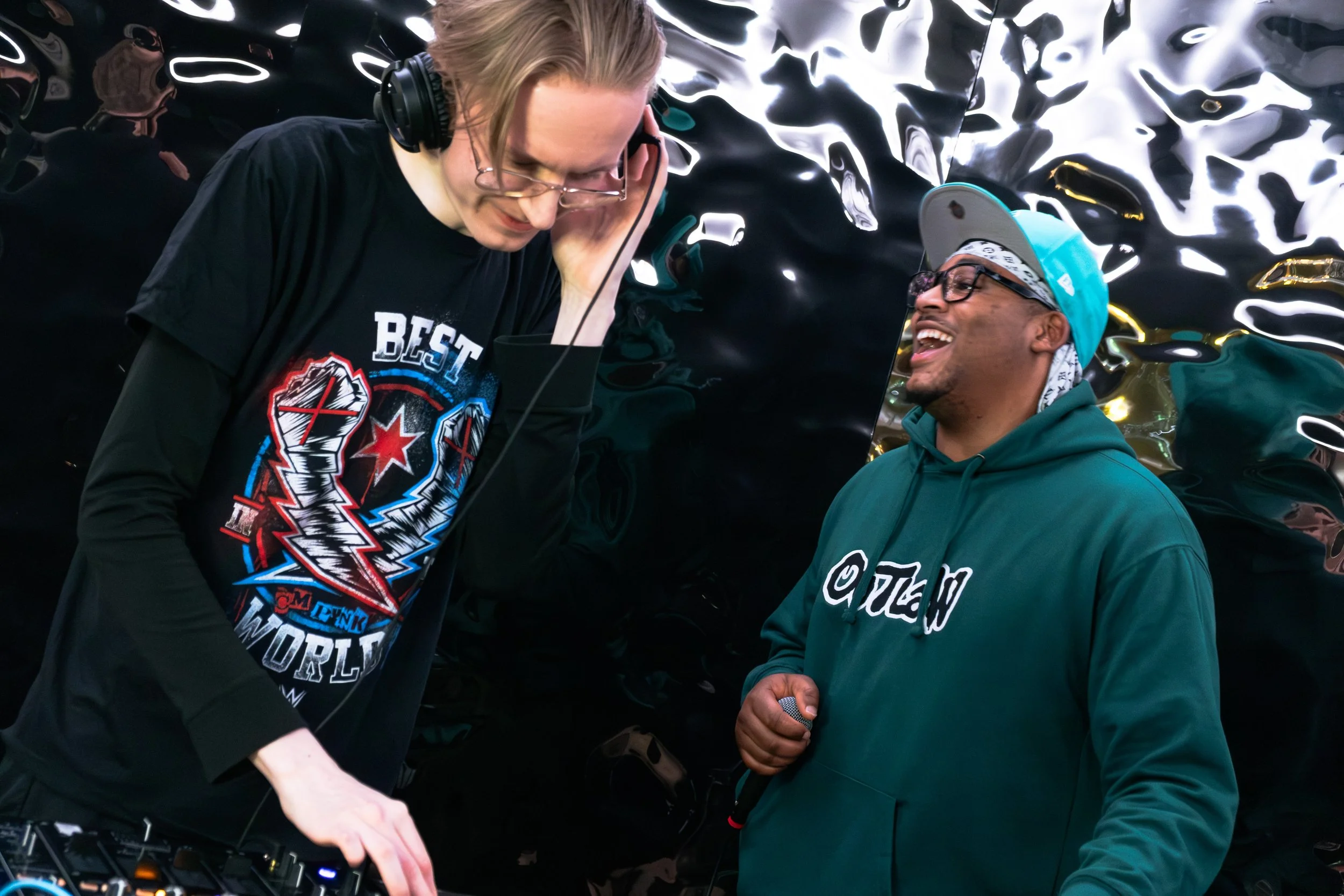 Two men are engaging in conversation at a DJ booth, one wearing headphones and a black shirt with colorful graphics, and the other wearing glasses, a teal hoodie, and a baseball cap, in front of a shiny, abstract metallic background.