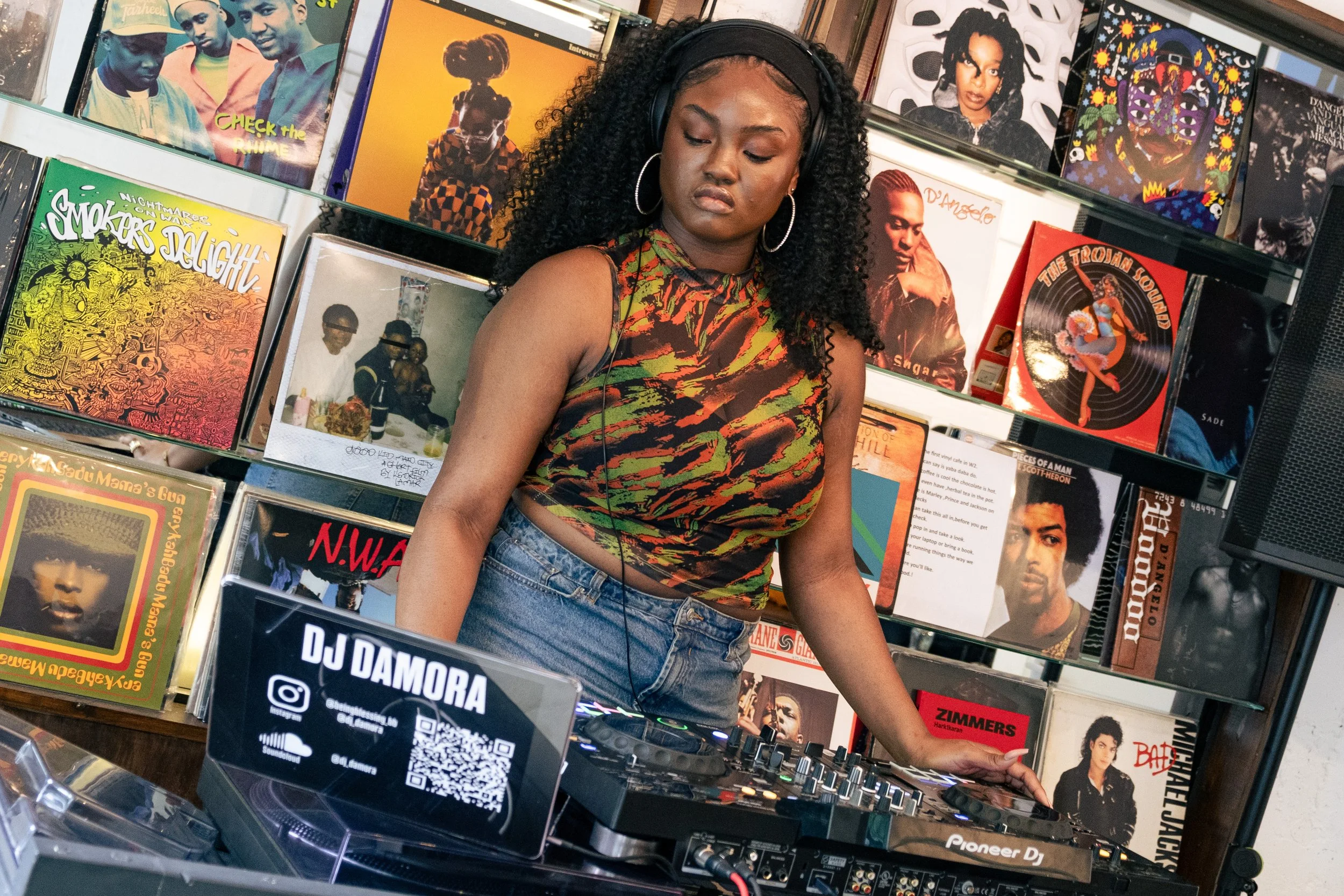 A woman with curly hair, wearing a colorful sleeveless top, DJing at a record store with vinyl albums displayed on shelves behind her. The albums include various artists and genres, and a DJ setup with a laptop and mixing equipment is visible in fron