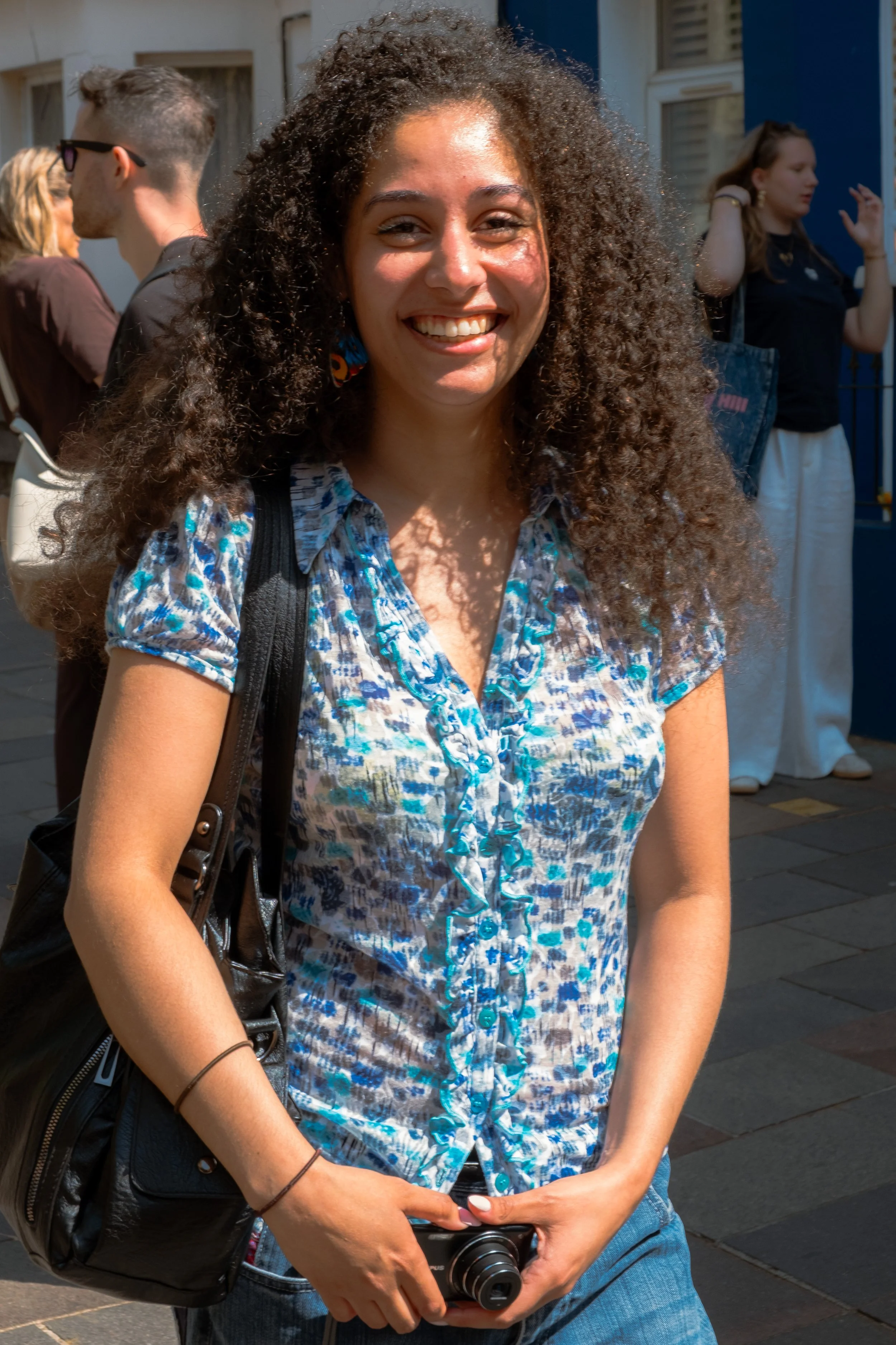 A woman with curly hair and a bright smile standing outdoors on a sunny day. She is holding a camera in her hands and wearing a blue patterned shirt. Other people are visible in the background, engaged in conversation.