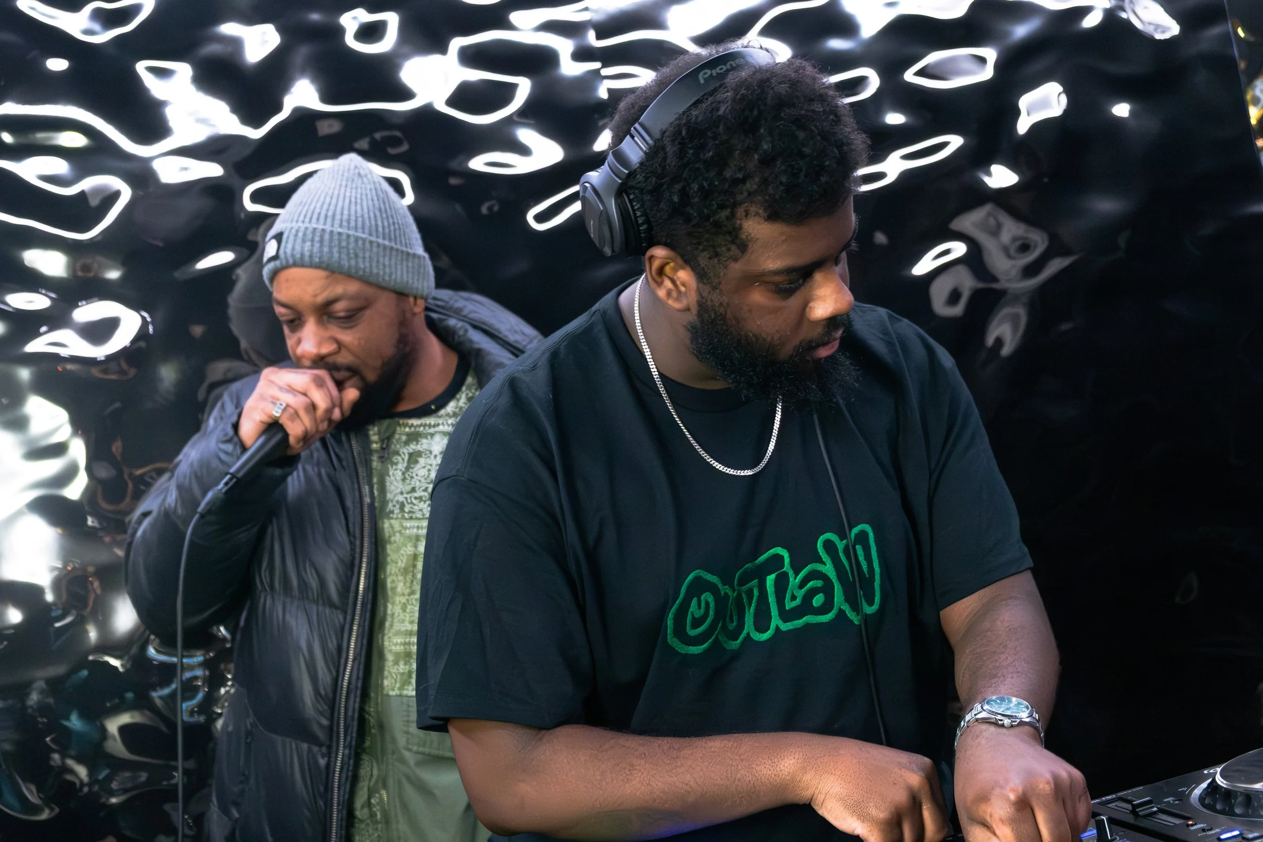 Two men with headphones and one with a microphone, standing close to each other against a black reflective background, possibly in a music or DJ setup.