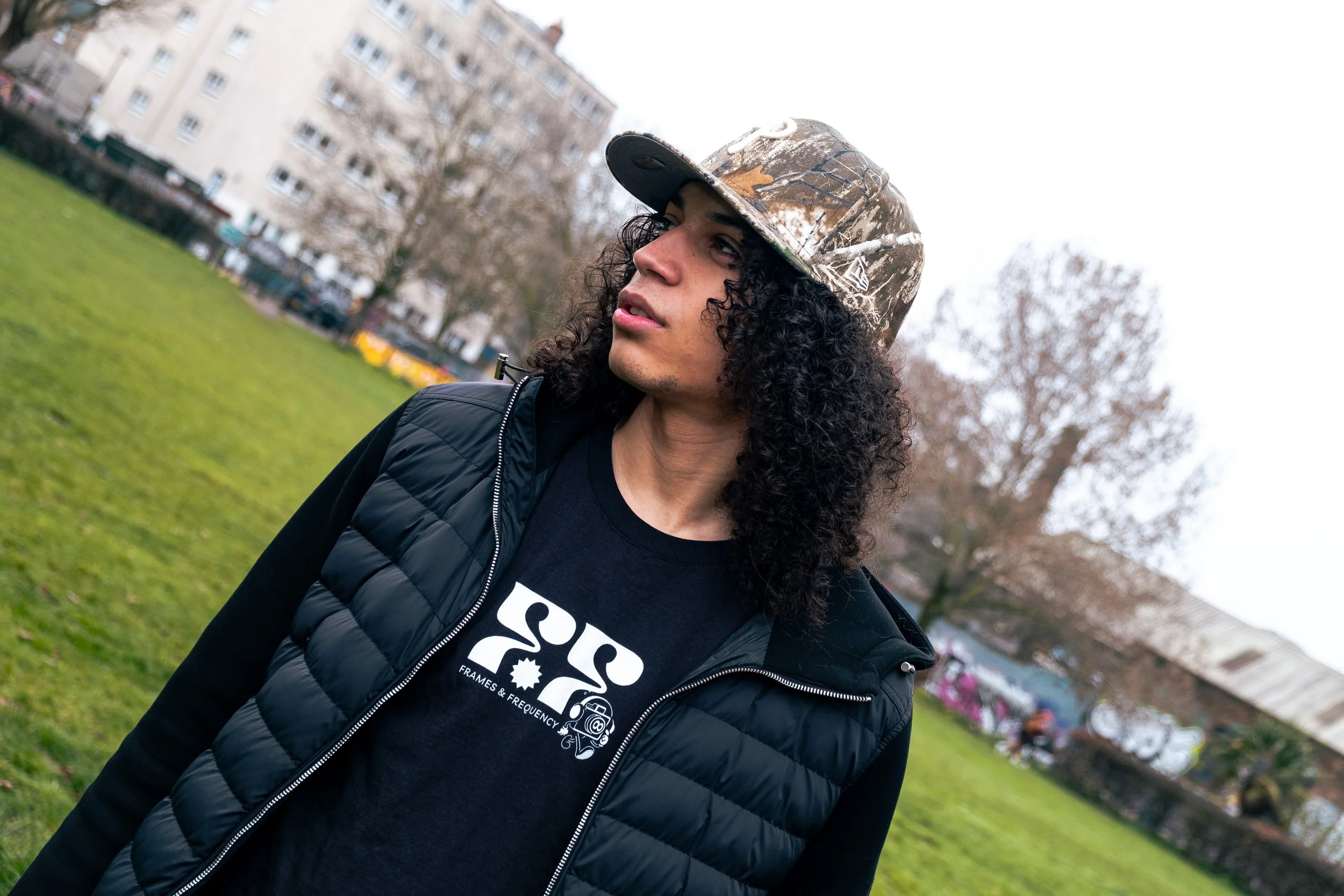 A young man with long curly hair wearing a camouflage baseball cap, black jacket, and black t-shirt with a logo, standing in a park with trees and buildings in the background on a cloudy day.