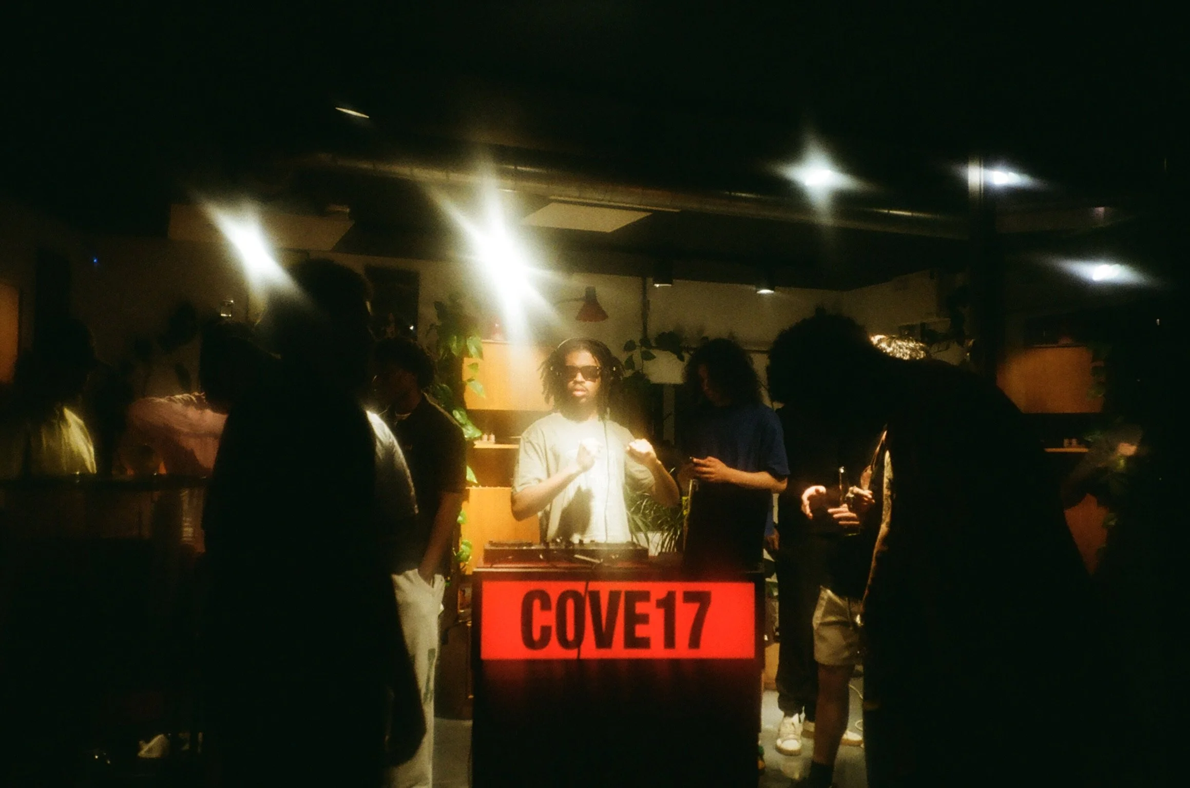 People gathered in a dimly lit indoor space, some wearing sunglasses, with a DJ or music controller in the center and a bright sign that reads ''COVERT'' in red letters.