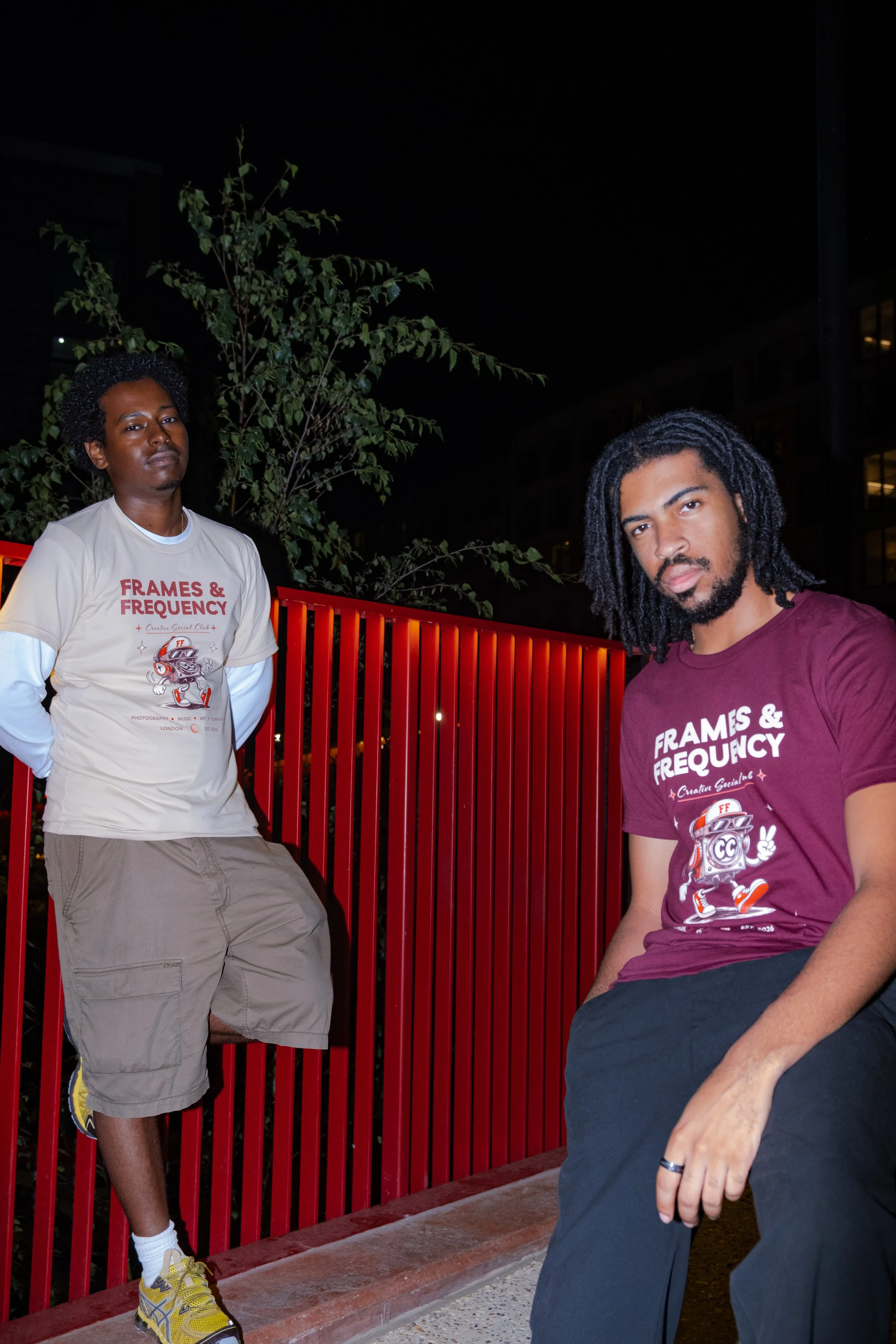 Two men wearing matching Frames & Frequency T-shirts pose at night beside a red railing and leafy tree.