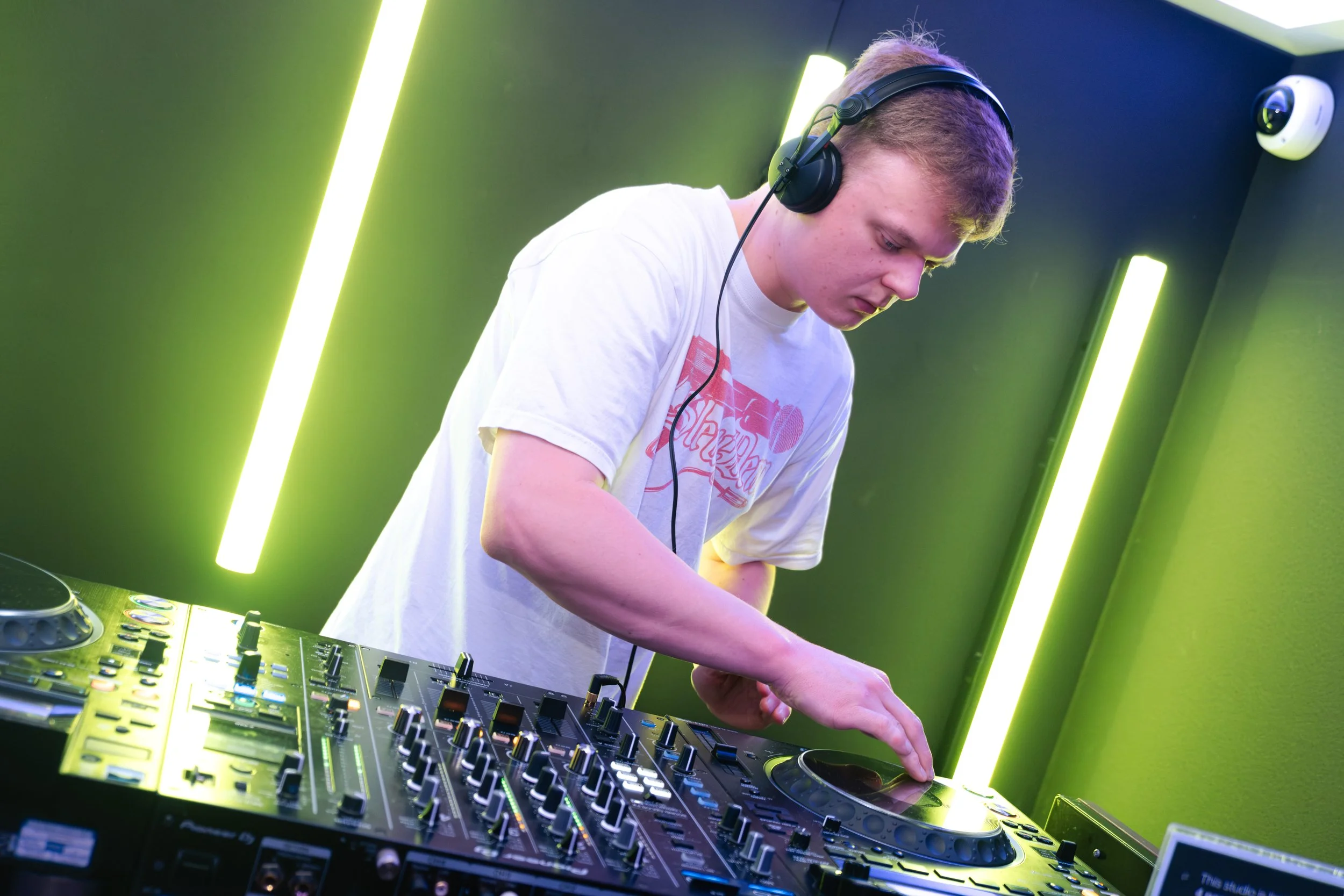 A young male DJ with blonde hair wearing headphones is focused on mixing music on a DJ console with numerous controls, illuminated by vibrant green and yellow lighting in a dark room.