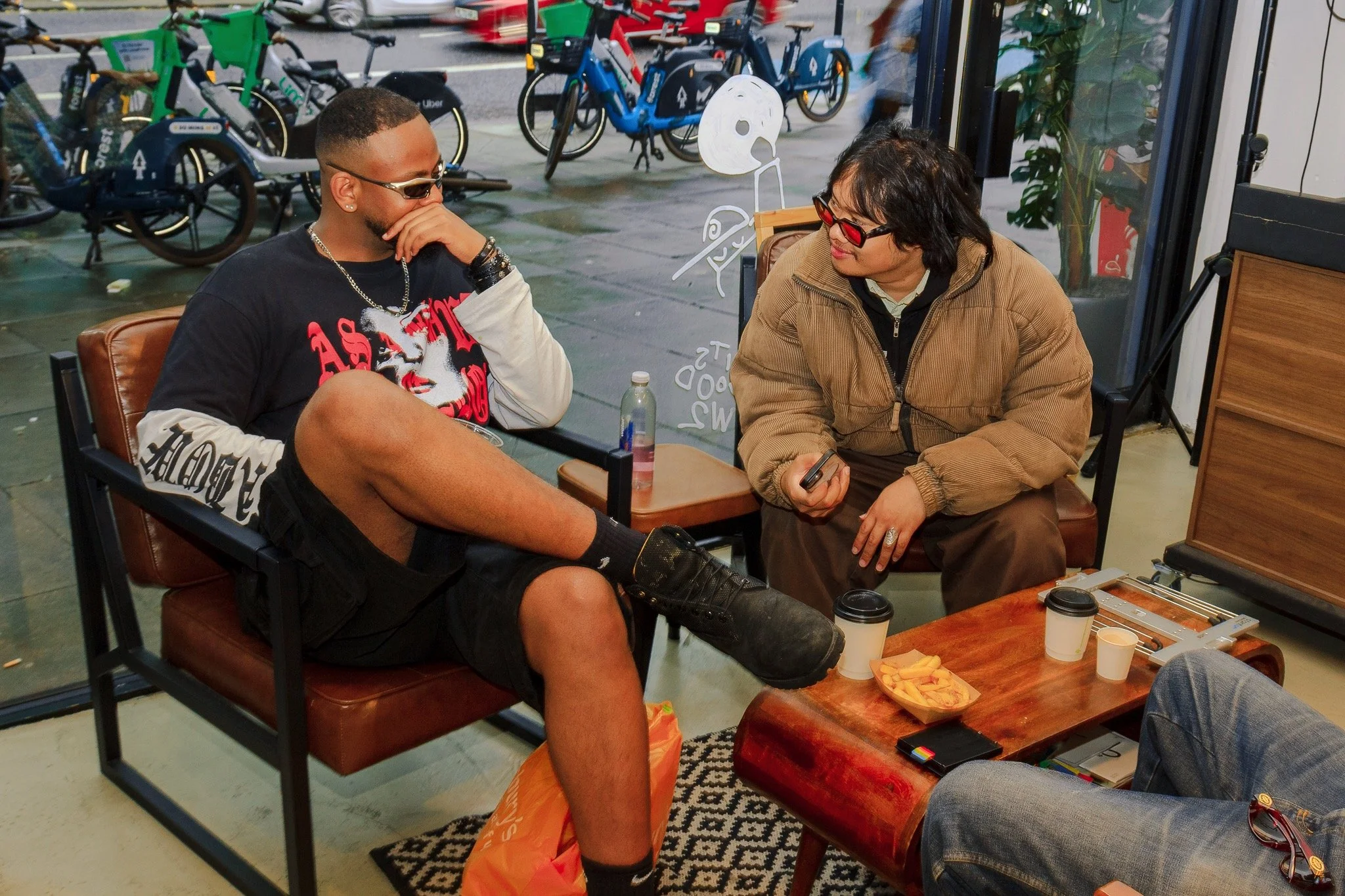 Two people sitting at a table in a cafe, engaged in conversation. The man on the left is wearing sunglasses and a black t-shirt with red and white graphics, and has a tattoo and jewelry. The woman on the right has short black hair, glasses, and is we