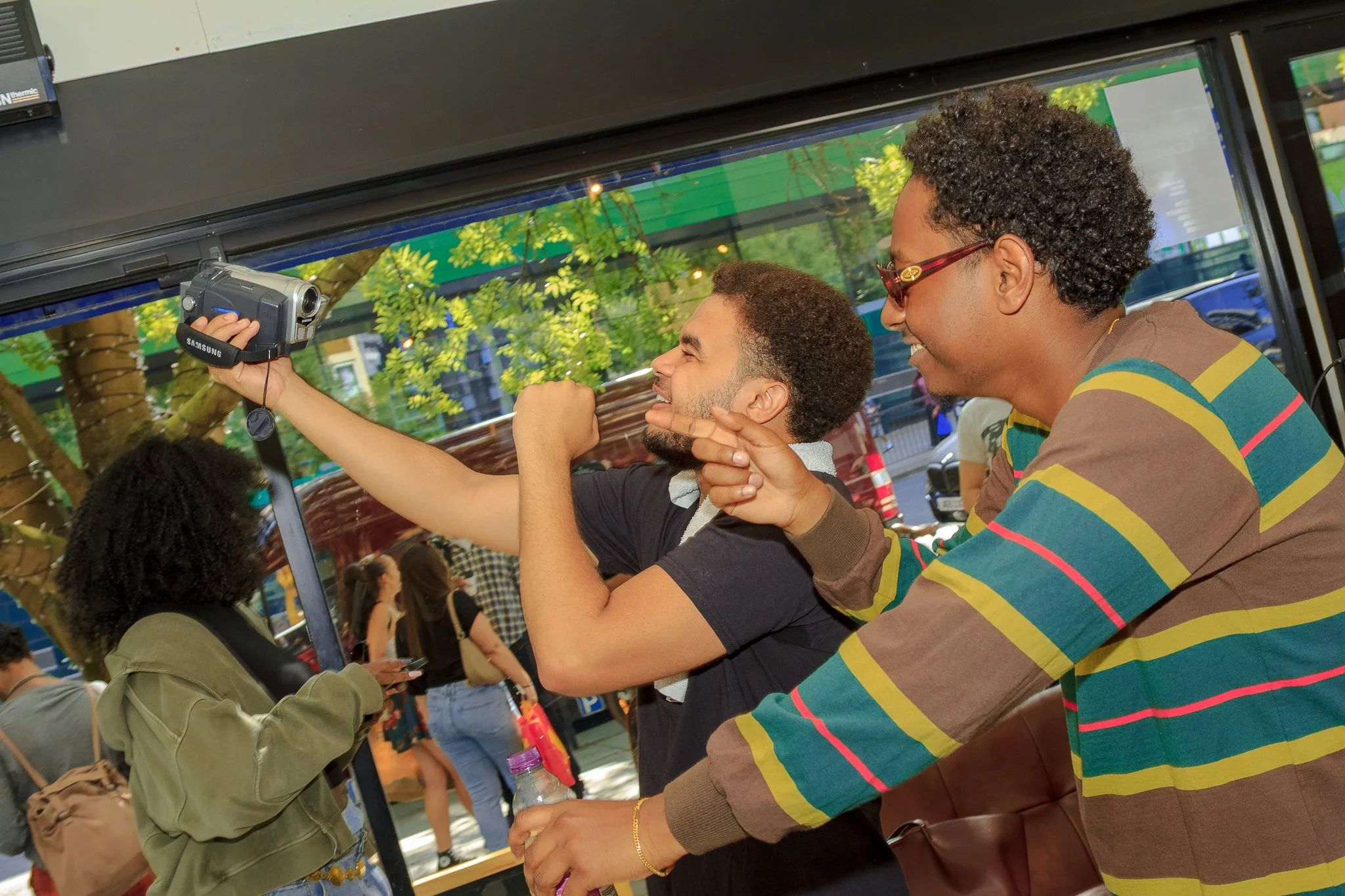 Two people taking a selfie inside a bus, with a woman smiling and wearing sunglasses, and a man making a gesture with his hand.