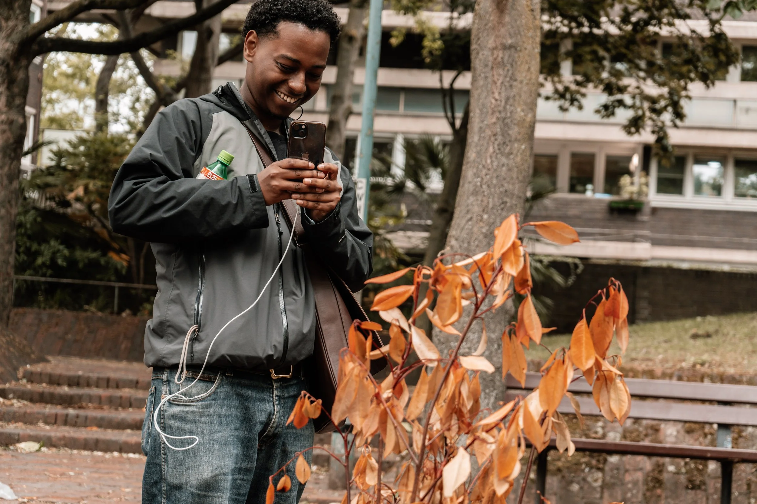 A young man outdoors smiling at his phone, wearing a black and gray jacket, standing near a small tree with orange leaves, with city buildings in the background.