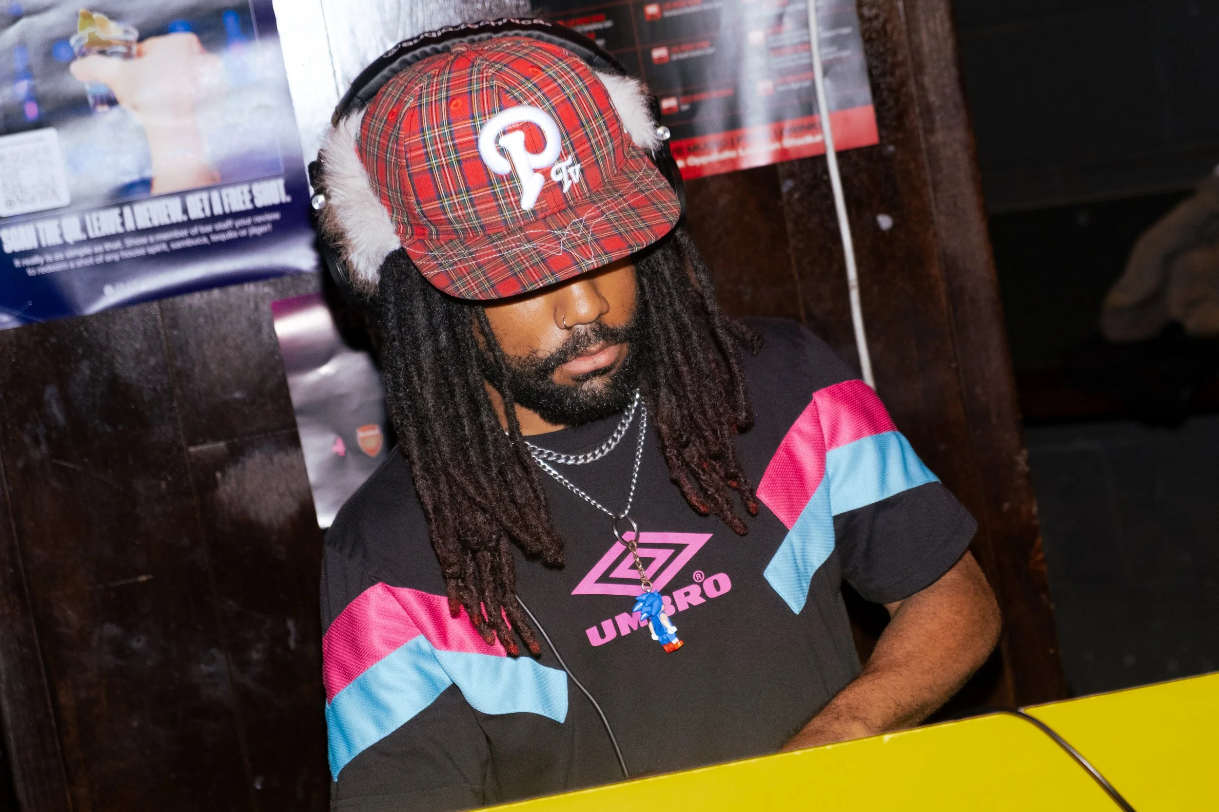 A person with dreadlocks wearing a red plaid hat with a headphone over it, a nose ring, and a colorful sports shirt, sitting at a table with yellow edges in a dimly lit indoor setting.