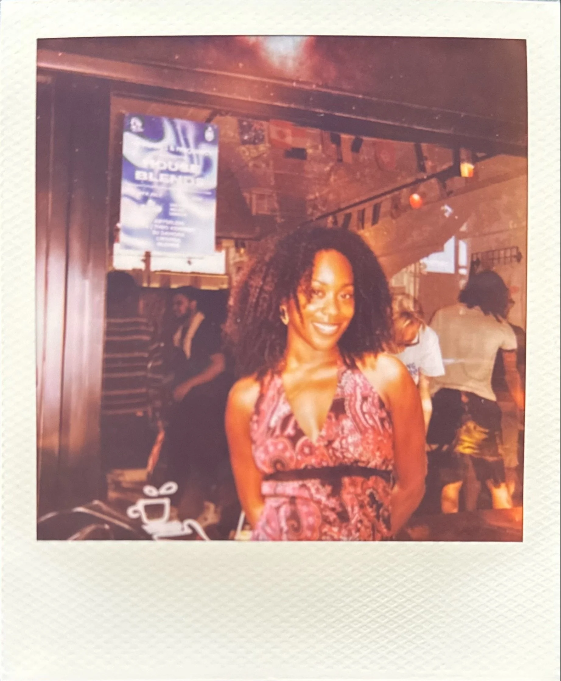 Smiling woman in a pink patterned dress posing inside a warmly lit restaurant or café