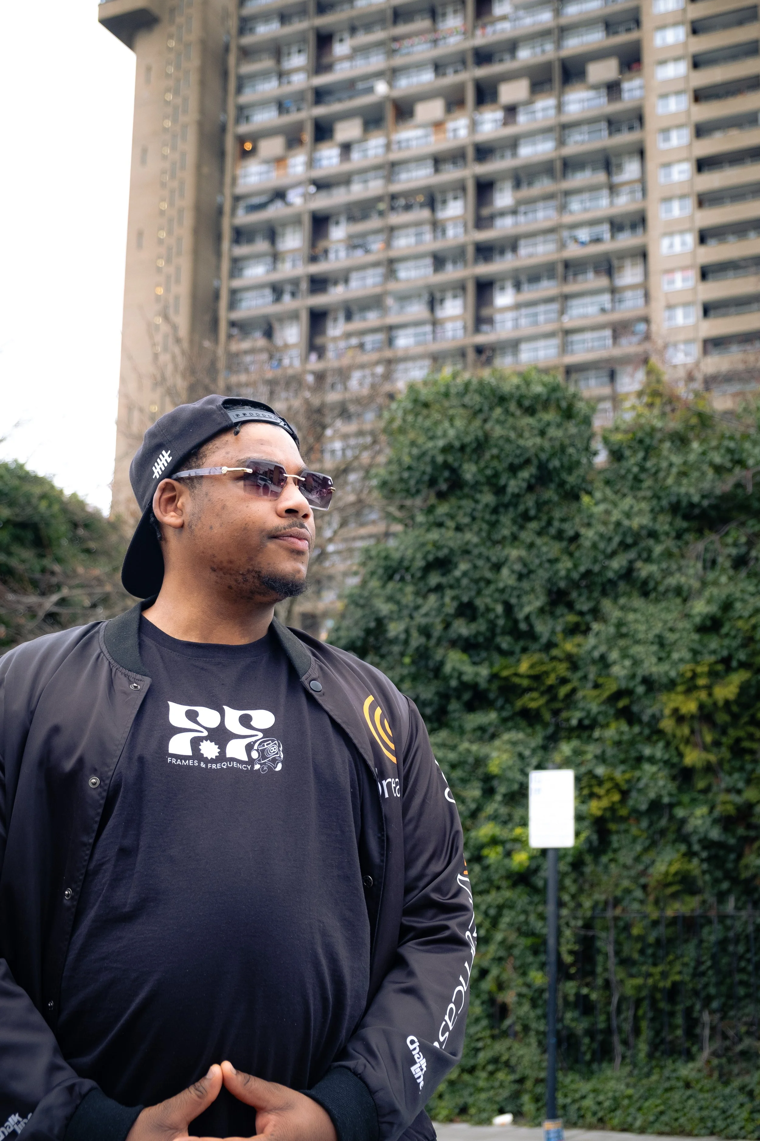 Man wearing sunglasses, black cap backwards, black jacket, and a T-shirt with graphic print, standing outdoors in front of a tall apartment building and green trees.