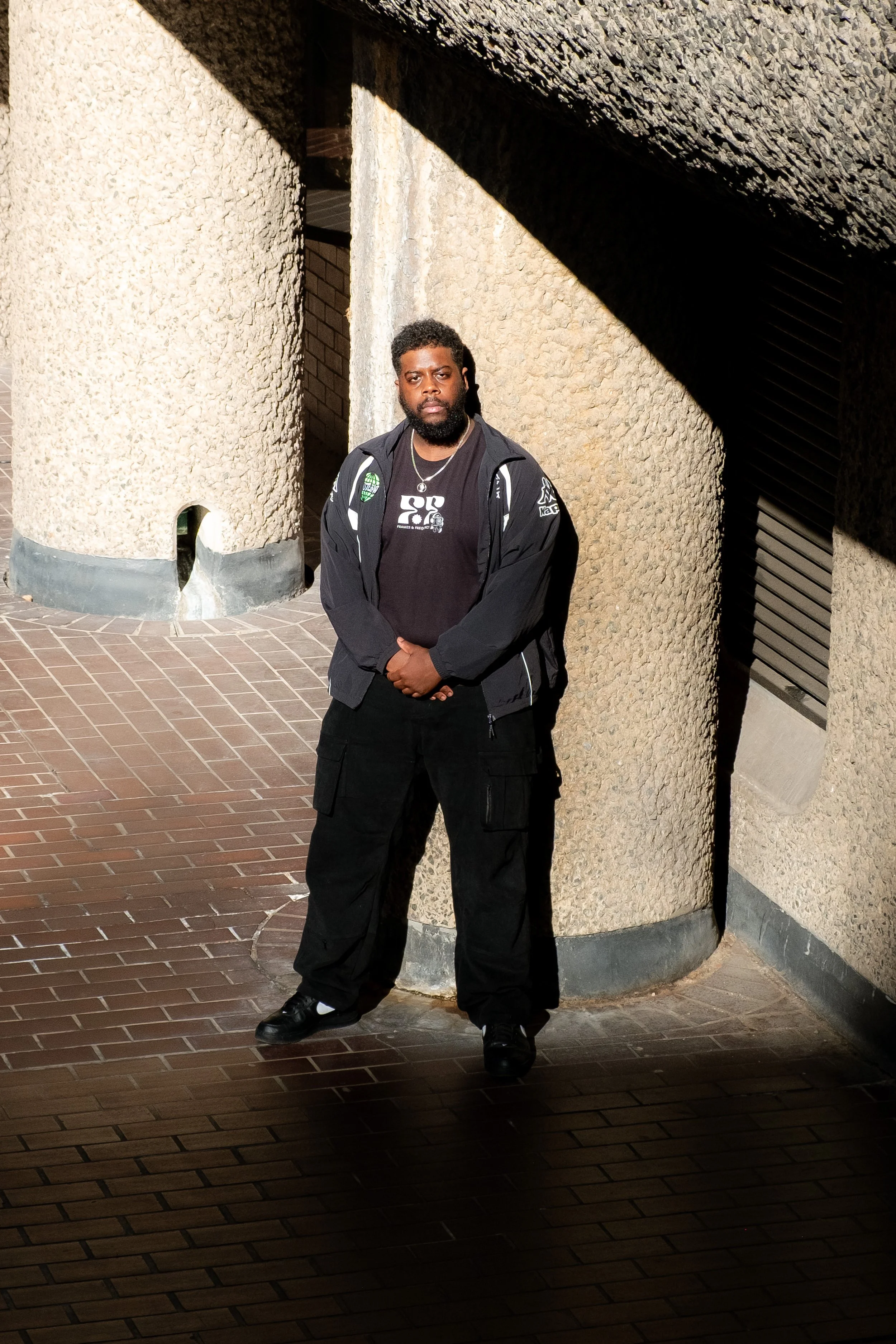 A man with dark skin and a beard standing outdoors against a textured beige wall. He is wearing a black jacket, black pants, and black shoes, with his hands clasped in front of him. Bright sunlight casts shadows on the wall and ground.