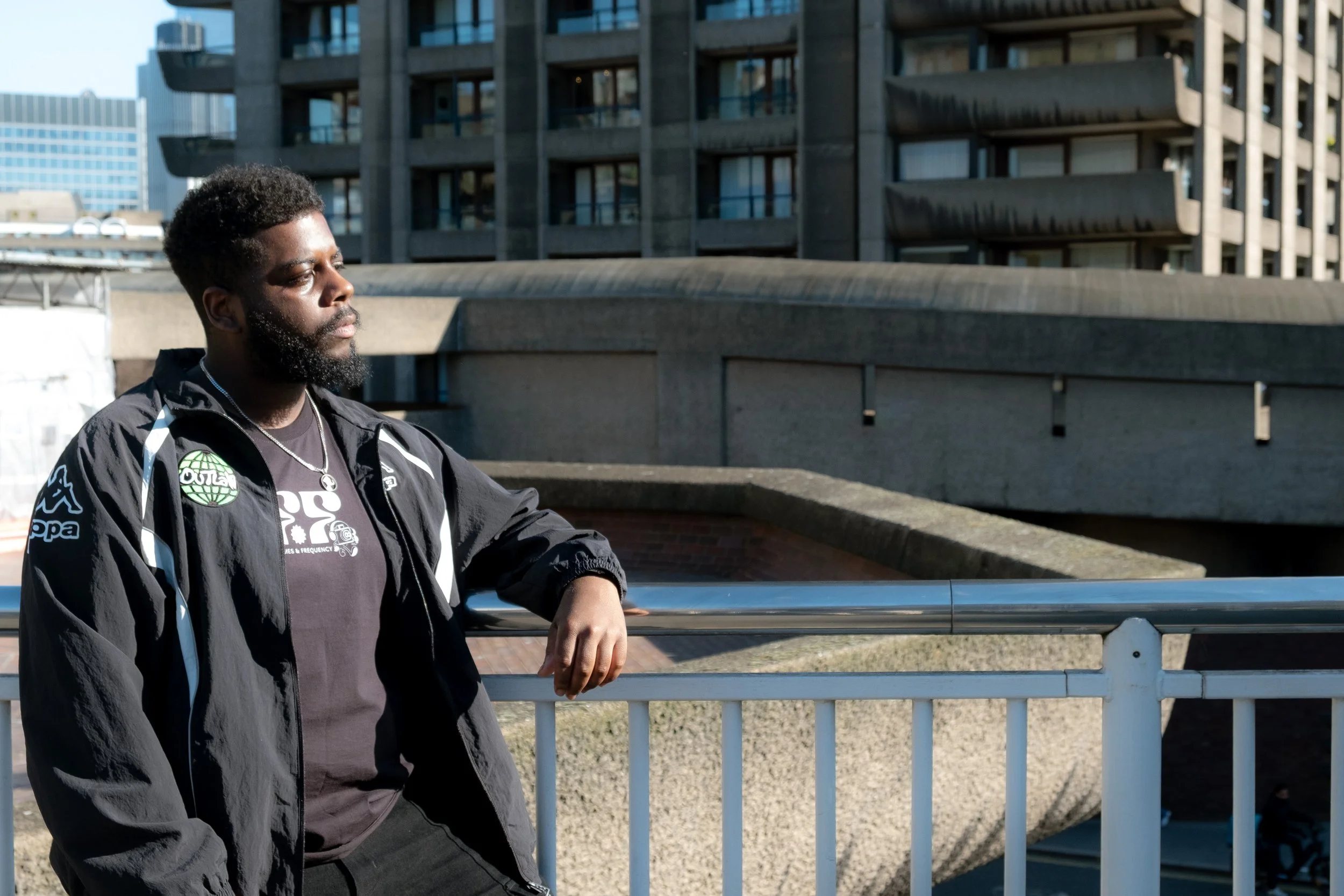 A young man with a beard and short hair standing outdoors near a metal railing with buildings in the background, wearing a black jacket and a graphic t-shirt.