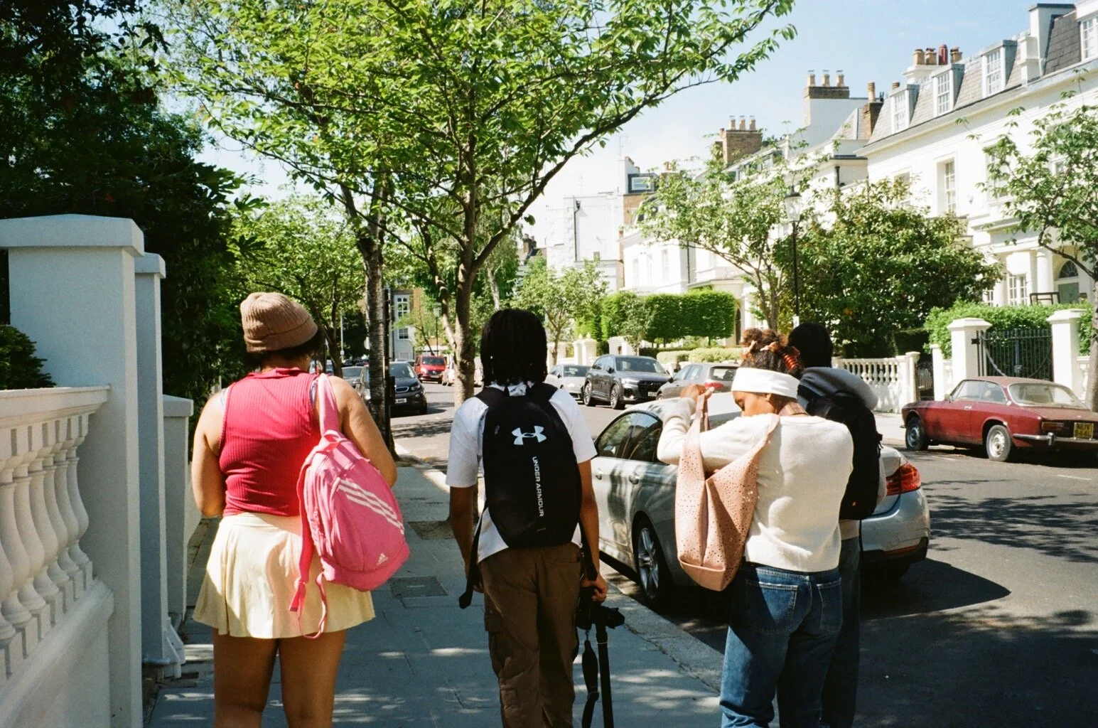People standing on a sidewalk in front of a row of parked cars, with trees and white buildings in the background on a sunny day.