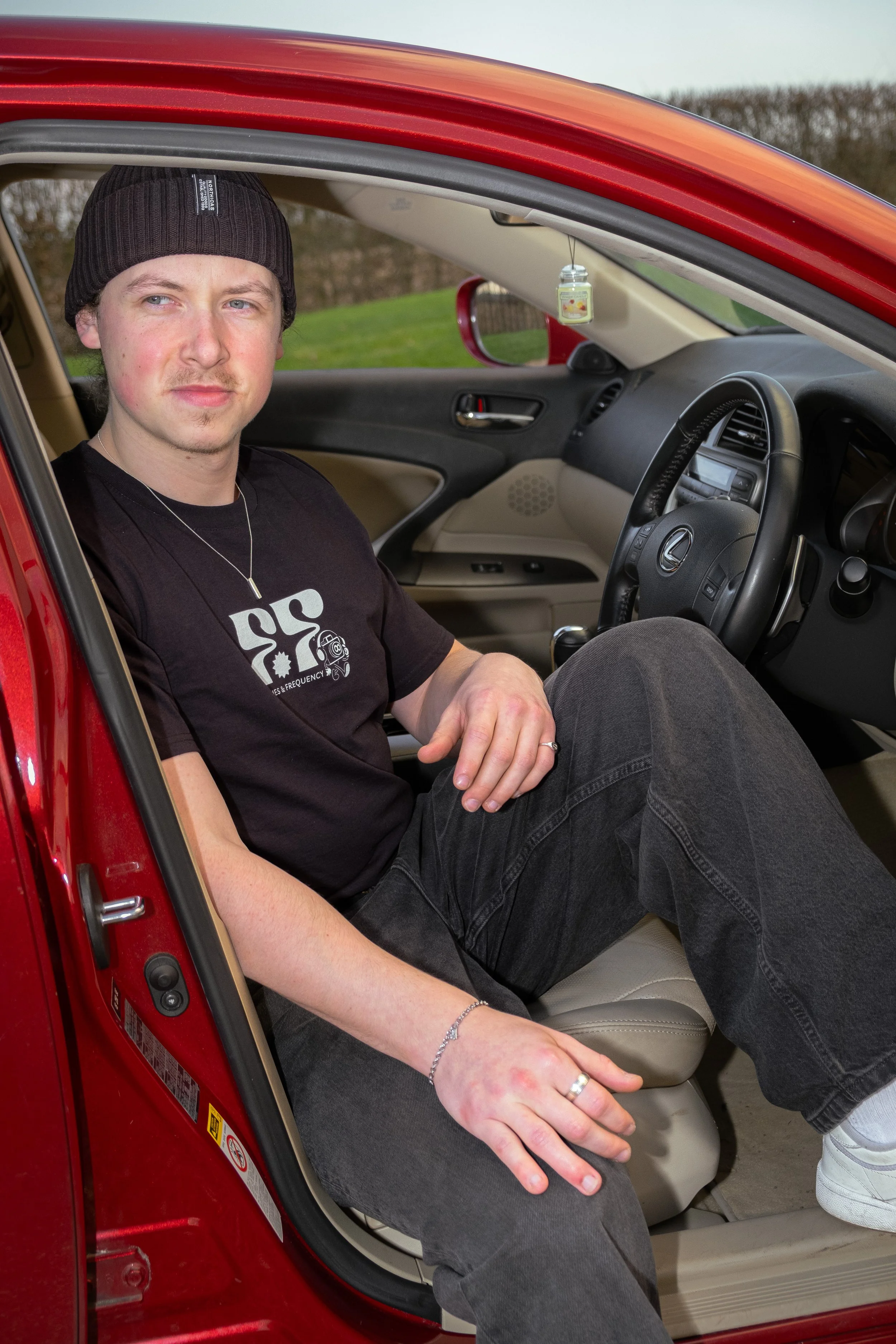 A young man sitting in the passenger seat of a red car, wearing a black beanie and a black T-shirt, with one arm resting on his leg. The car's interior is beige, and the background shows an outdoor setting with trees and grass.