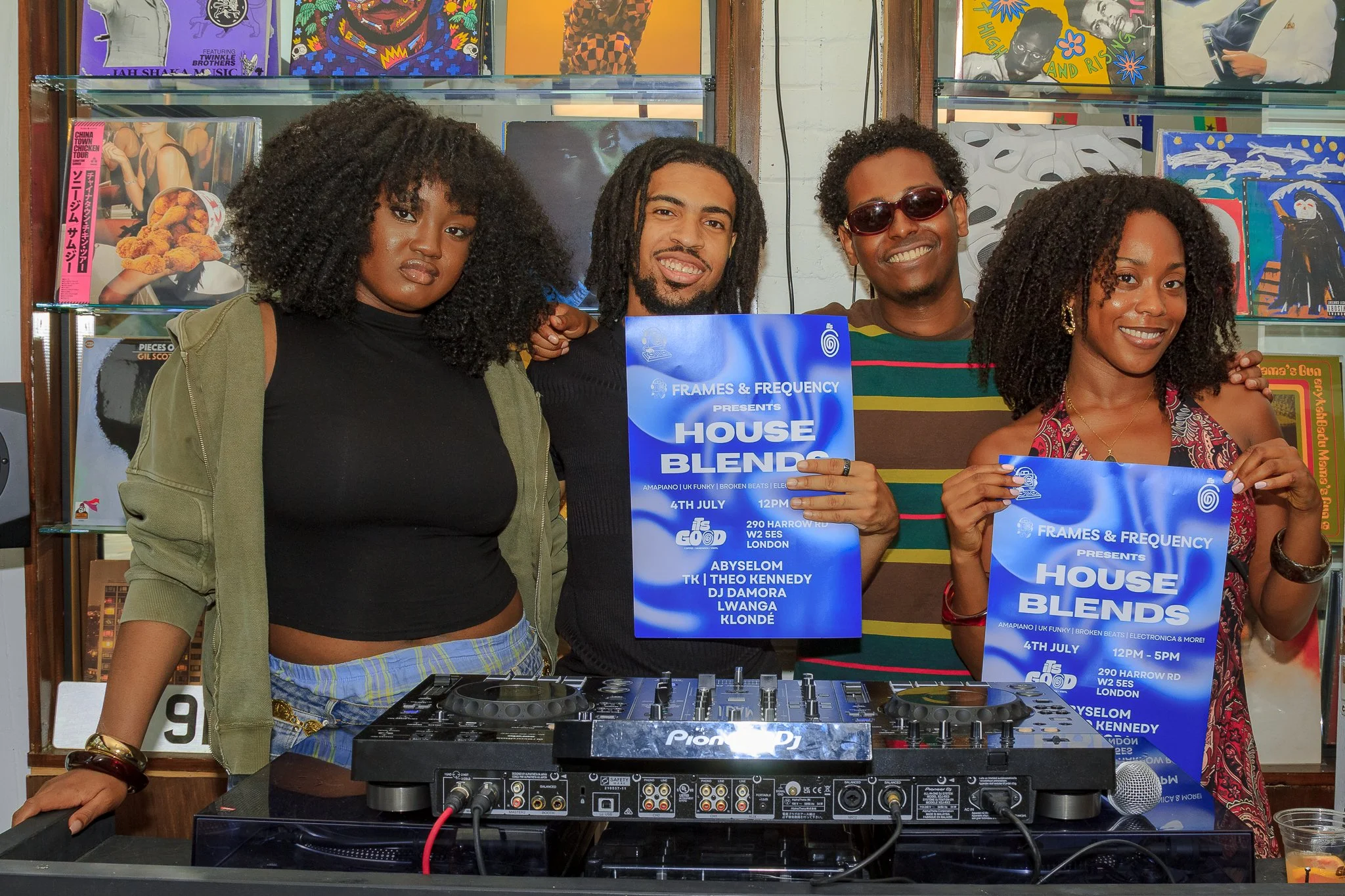 A group of five diverse people standing behind a DJ booth at an event, holding flyers for a house music event called 'House Blends' scheduled for July 4th. They are smiling and dressed casually, with a backdrop of colorful pop art and record covers.