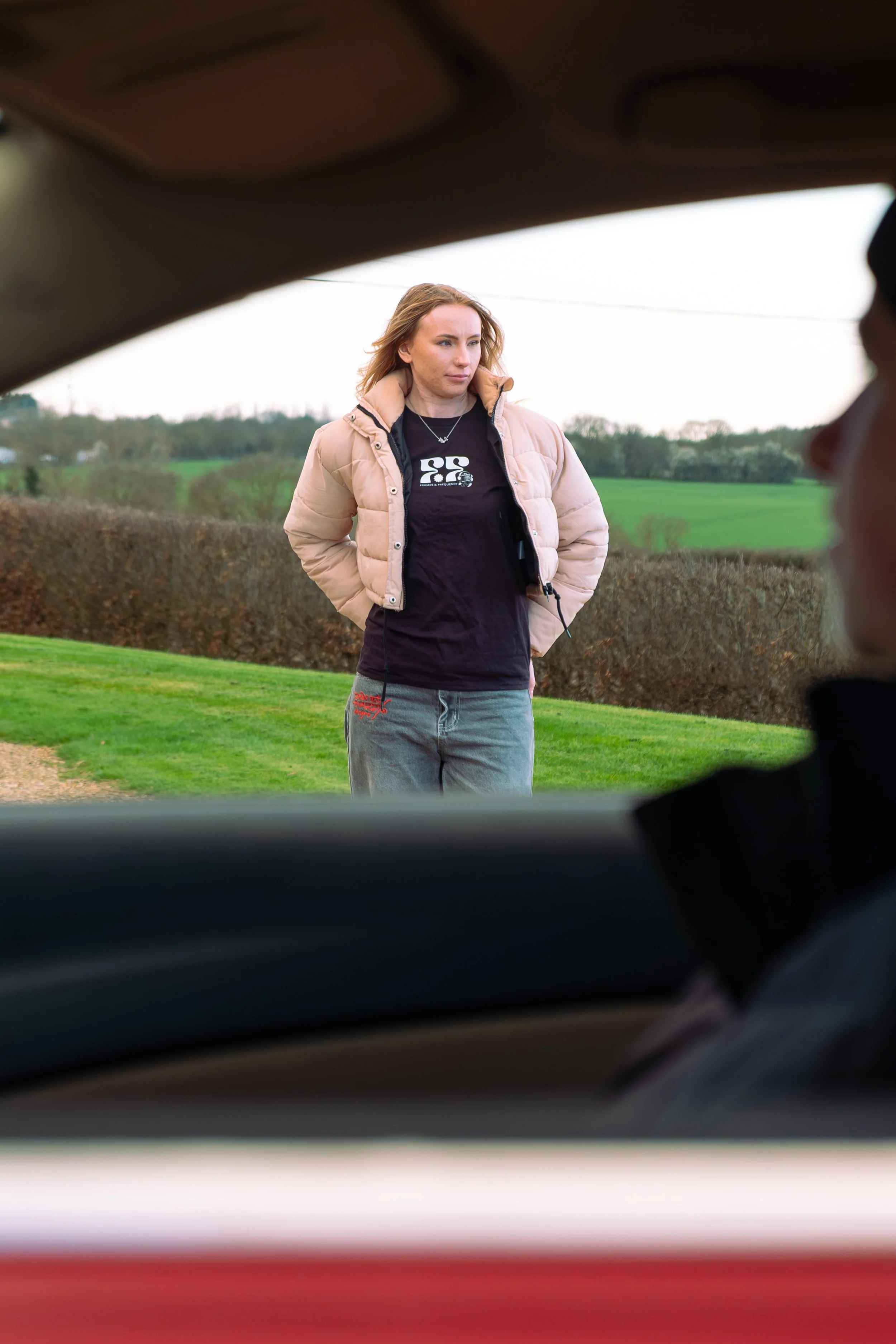 A woman standing outdoors, wearing a beige jacket and a black shirt, looking off to the side. The image is taken from inside a vehicle, with the frame of the car window surrounding her.