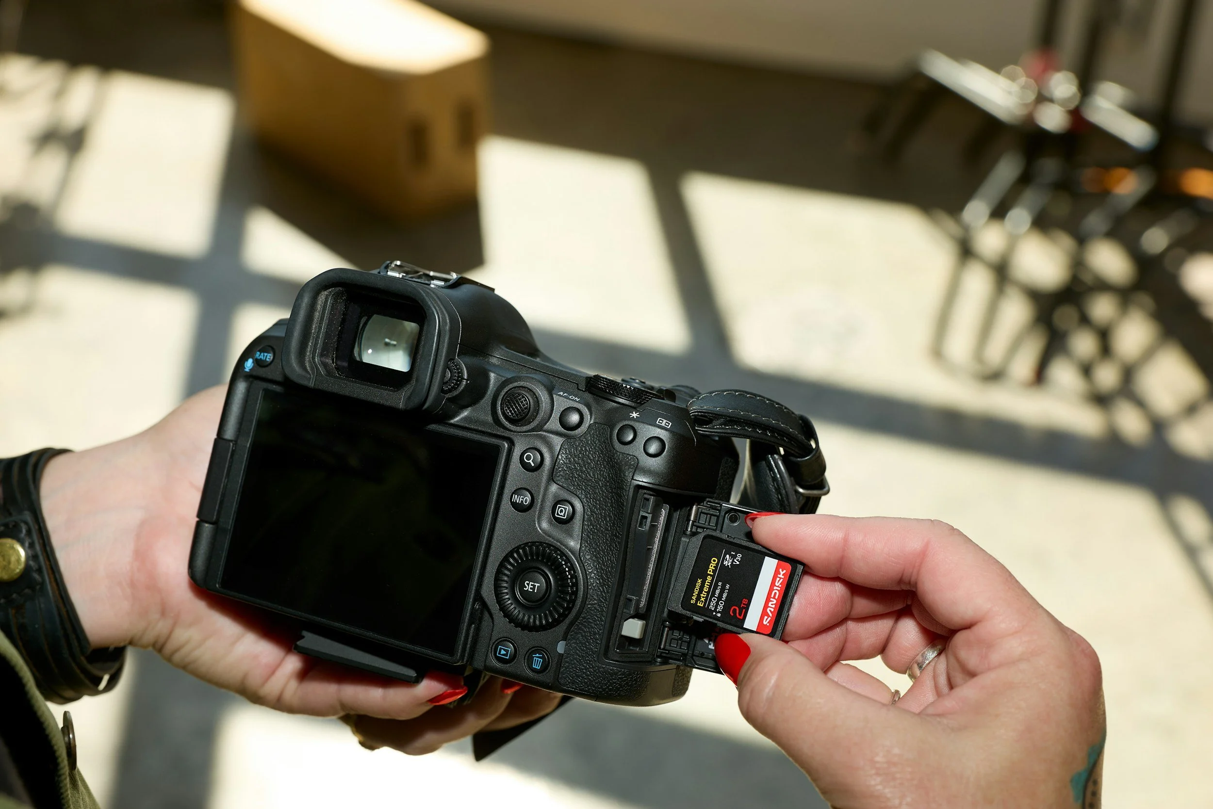 Person inserting a memory card into a digital camera, with a blurred background of furniture and sunlight.