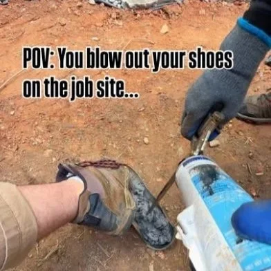 A person wearing gloves holds a gas canister while another person attempts to blow out a pair of work shoes on a dirt surface.