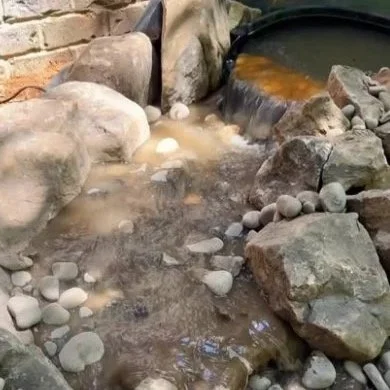 A small artificial pond with rocks around it, filled with muddy water, and a bubbling filter or waterfall feature enclosed by stones.