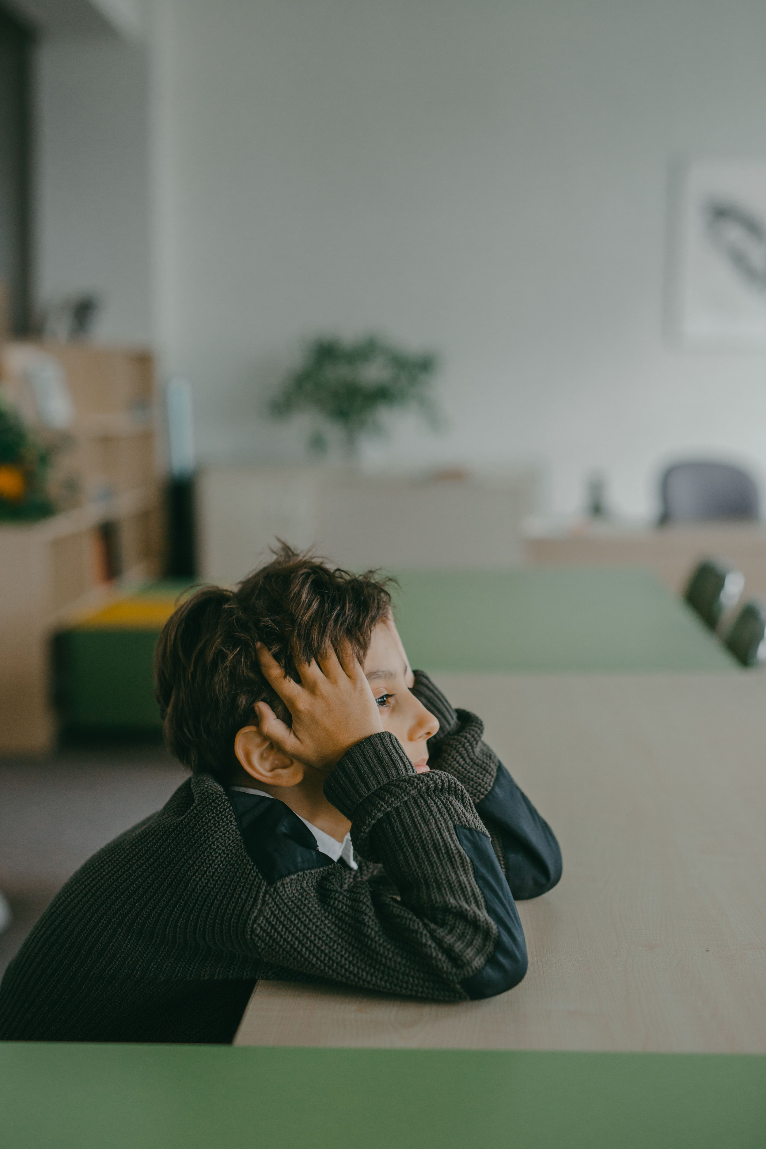 A young boy with brown hair sits at a desk, resting his head in his hands, looking pensive or distracted.