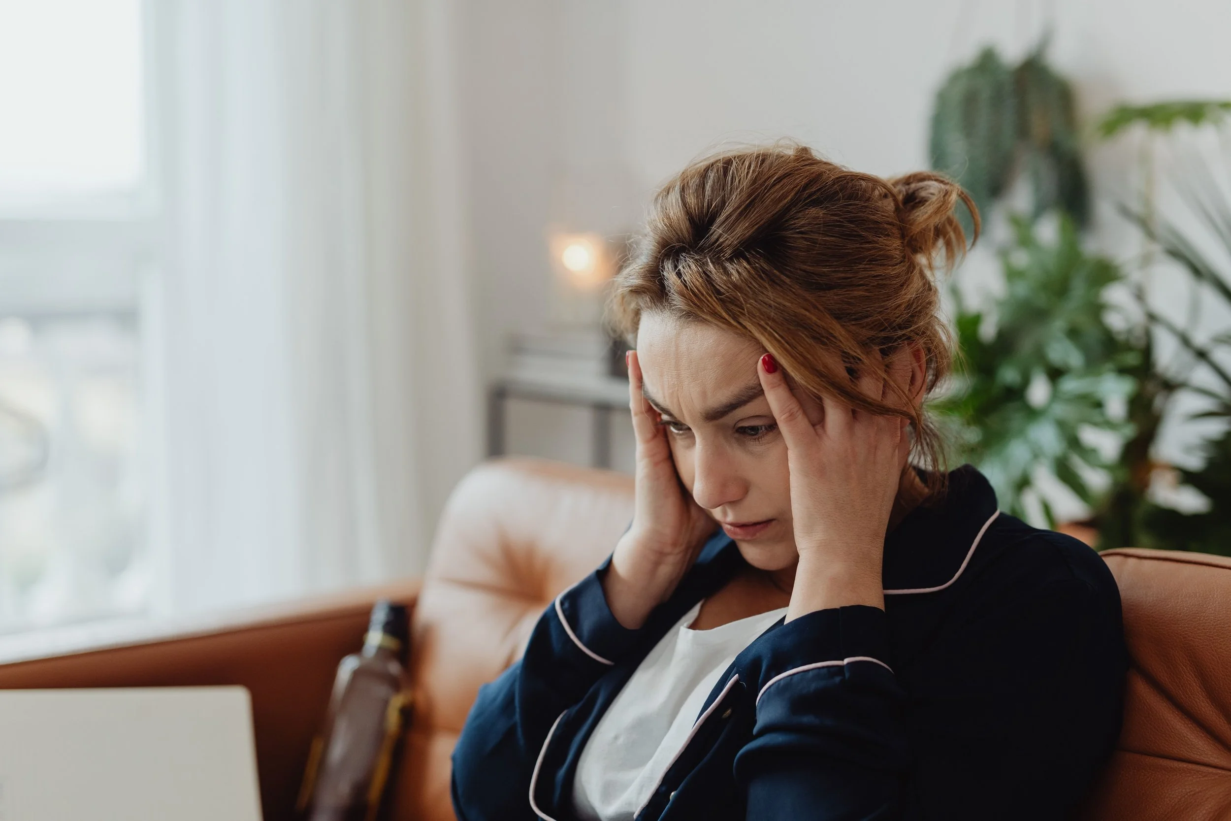 A woman with reddish-brown hair and red nail polish, wearing a navy blue sweatshirt with white piping and a white shirt underneath, sitting on a tan couch with her hands on her temples in a distressed or tired gesture.
