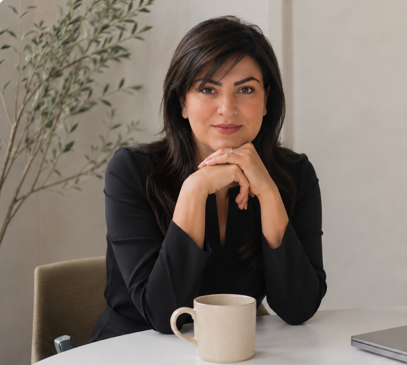 A woman with dark hair and a black blazer sitting at a table with her hands under her chin, looking at the camera. There is a beige mug and a closed laptop on the table, and a plant in the background.
