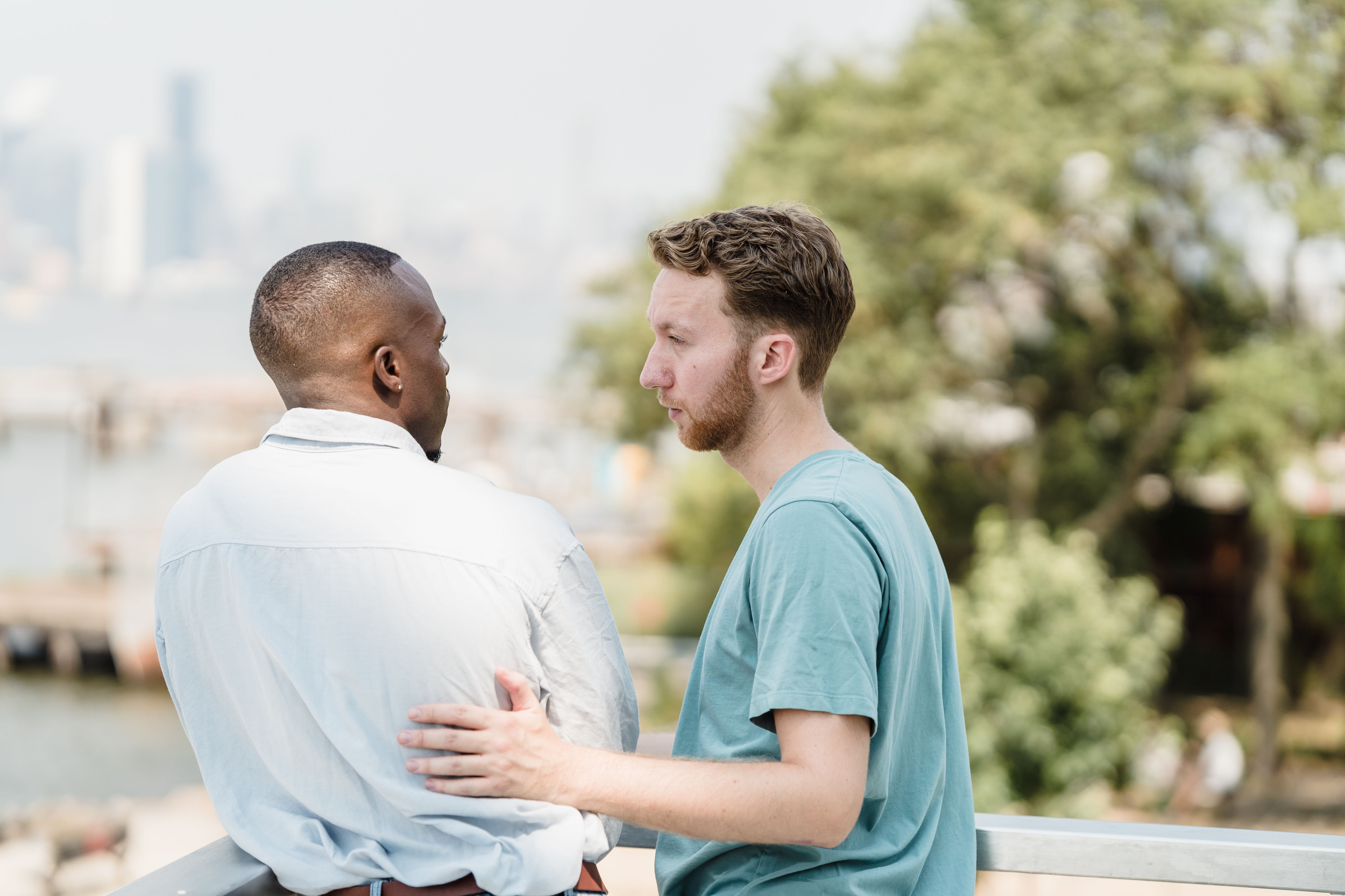Two men are facing each other closely, one with dark skin and short hair and the other with light skin and curly hair; they are outdoors near a railing, with trees and a cityscape in the background.