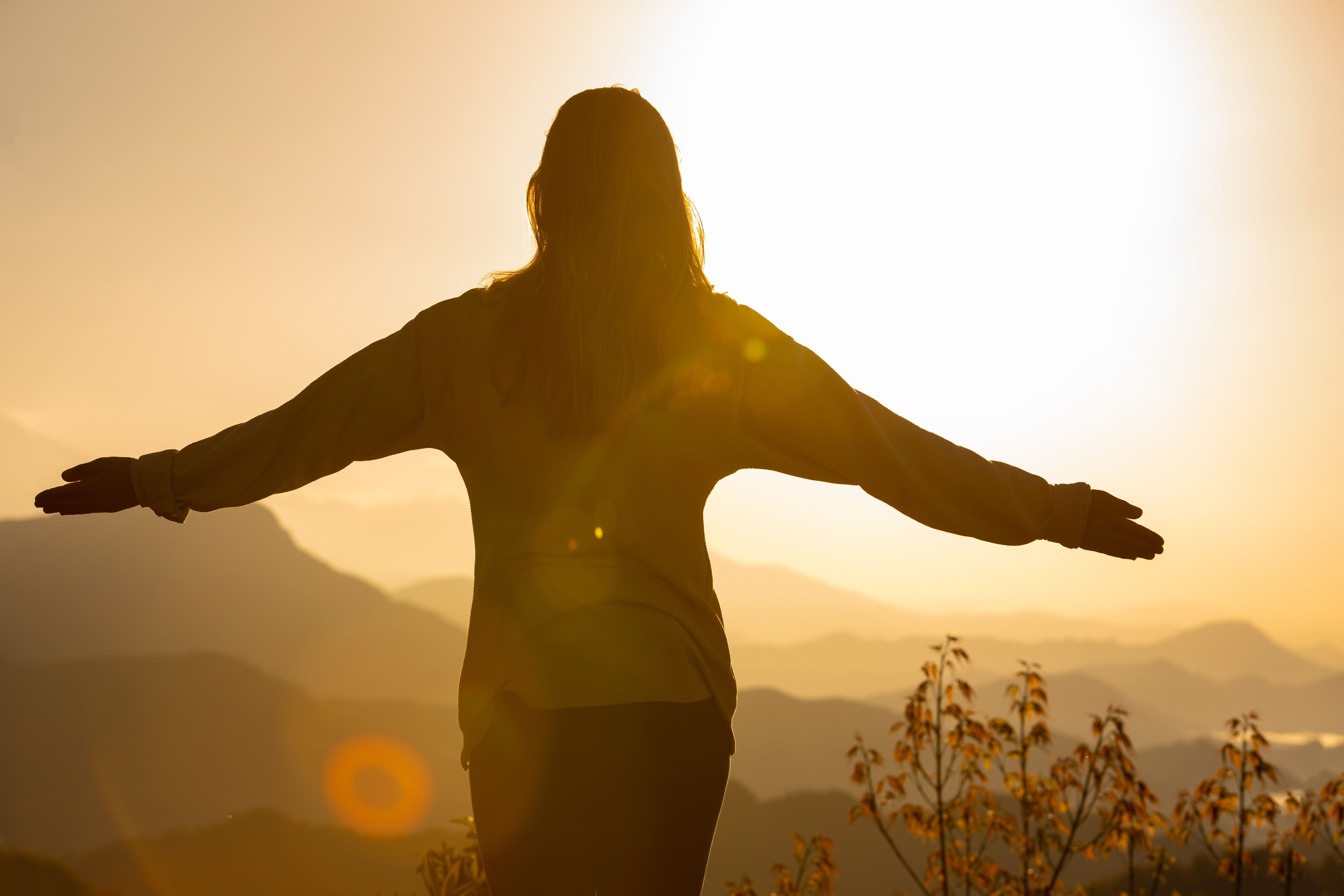 Person with arms outstretched at sunset or sunrise with mountains in the background.