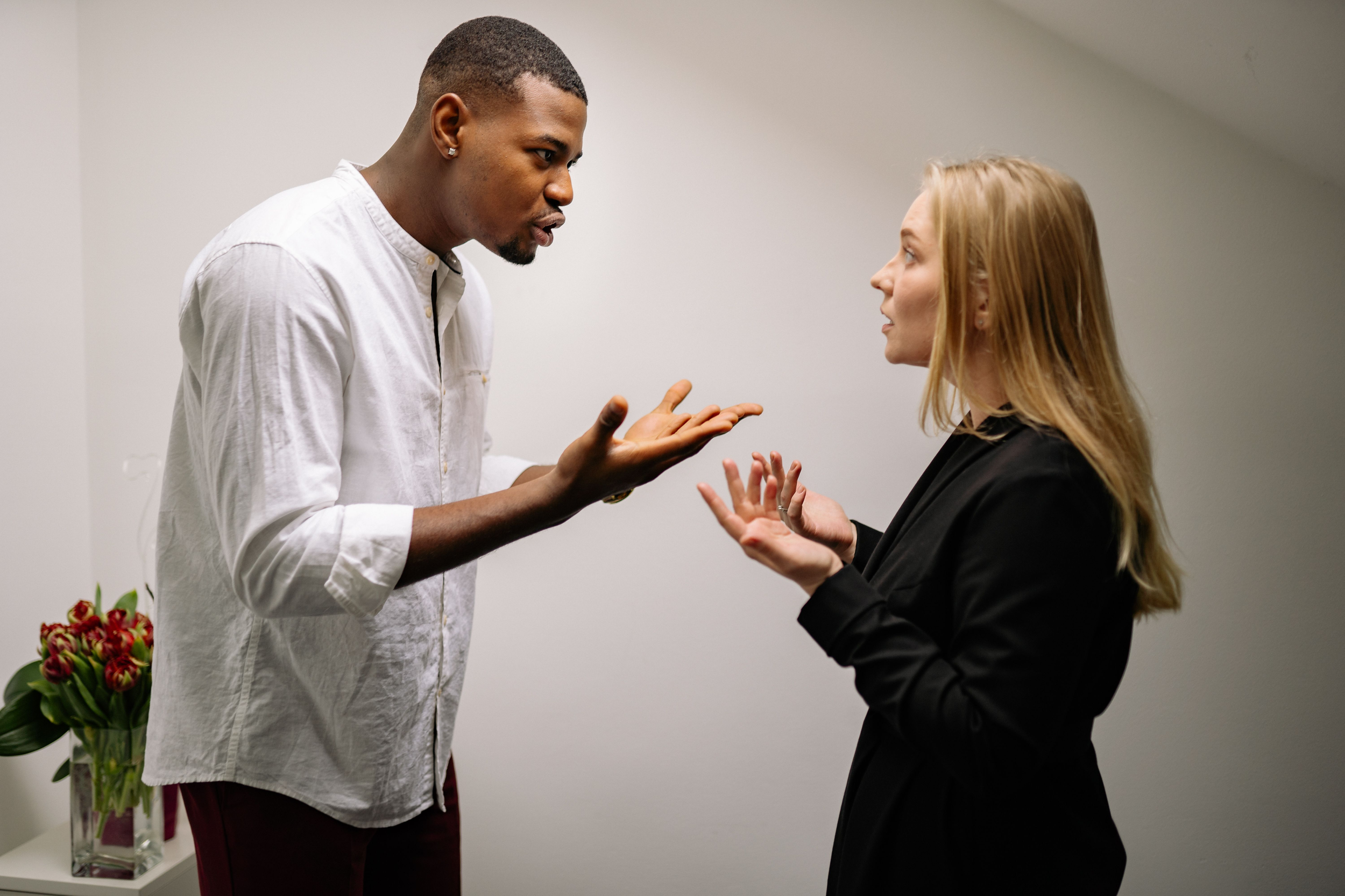 A man and woman are having a serious conversation indoors. The man is gesturing with his hands while the woman listens attentively. There is a vase of flowers on a table in the background.