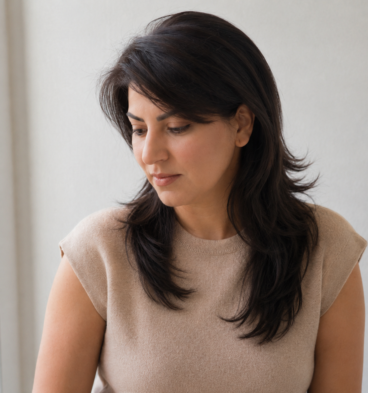 A woman with shoulder-length dark hair and light skin, wearing a beige short-sleeved top, looking down thoughtfully against a plain light-colored wall.