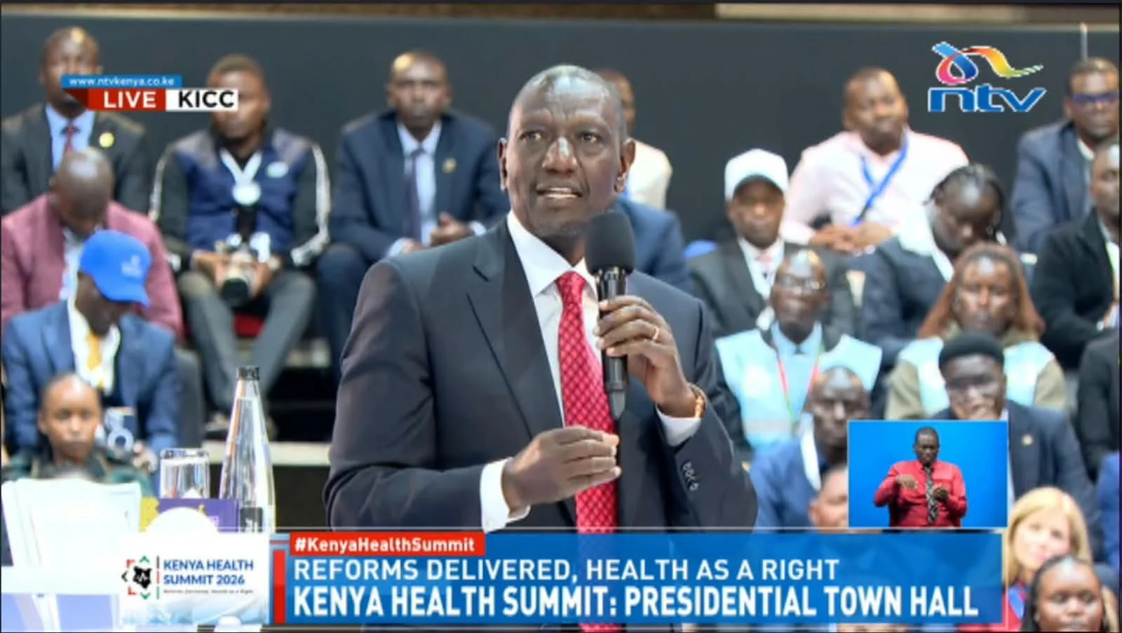 President William Ruto in a dark suit and red tie speaks into a microphone at a podium, with a seated audience behind him at the Kenya Health Summit town hall event; text overlays read “Reforms Delivered, Health as a Right” and “Kenya Health Summit.”