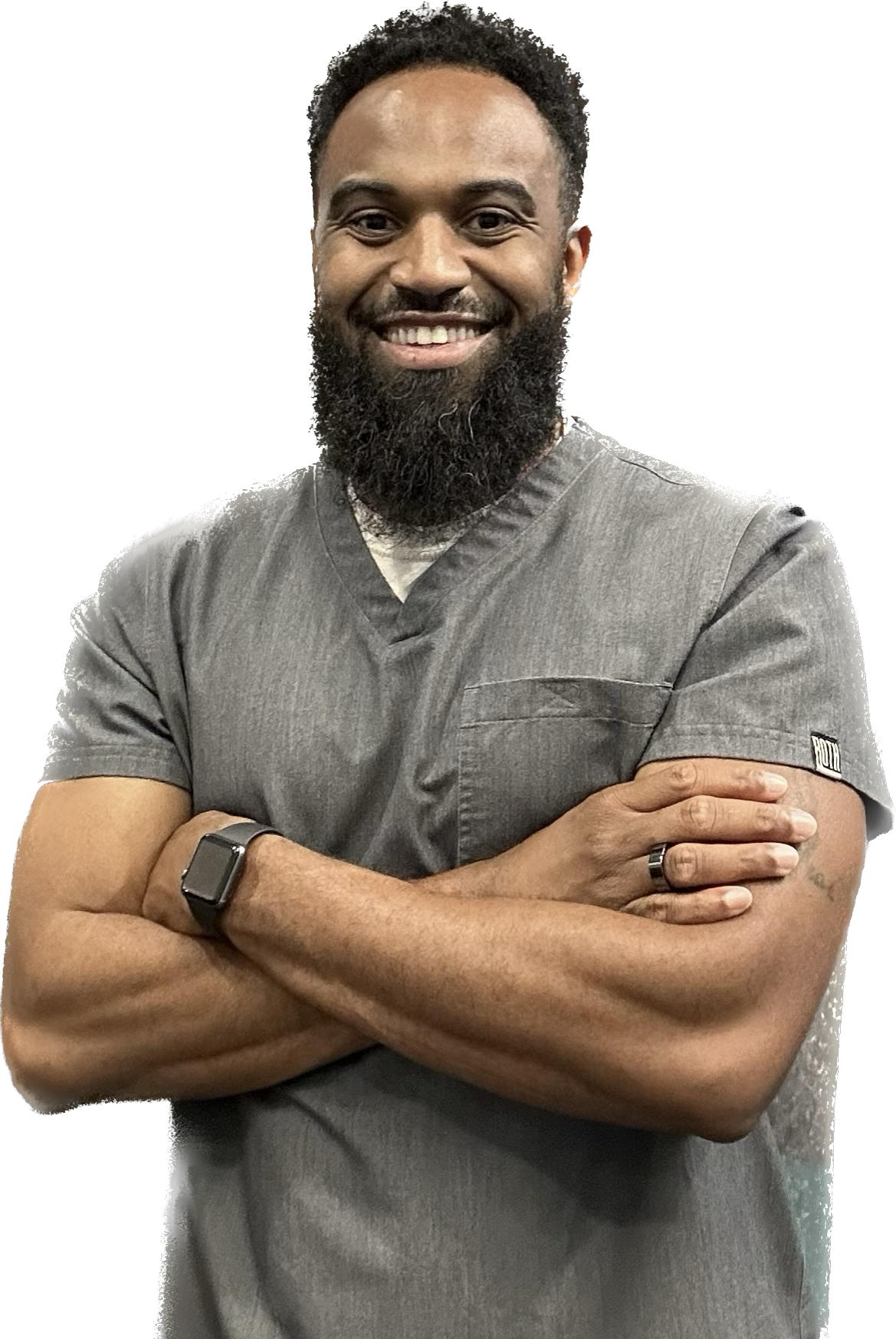 A smiling man with a beard and short curly hair wearing gray scrubs, arms crossed, with a smartwatch on his left wrist, standing against a plain background.