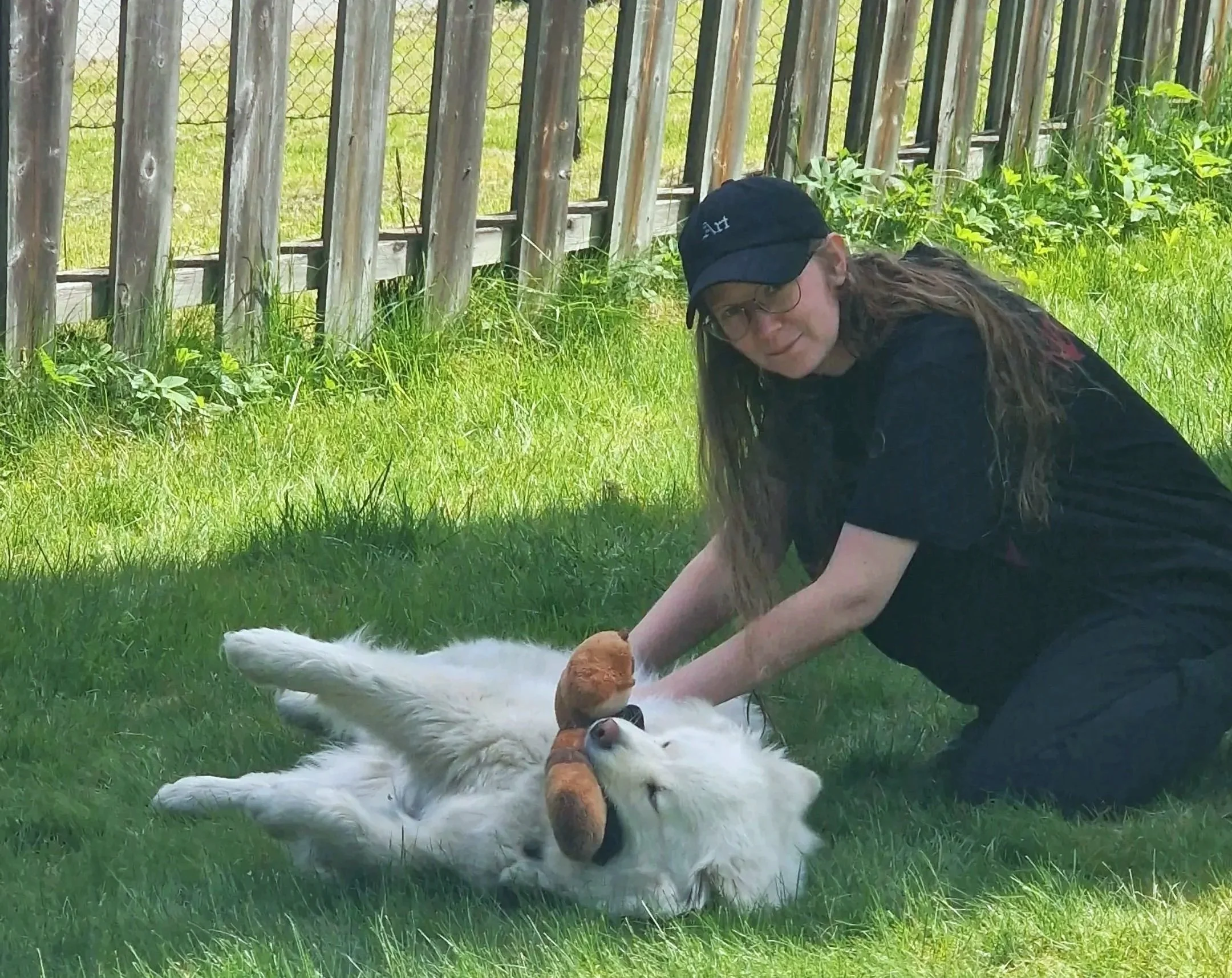 A woman in a black cap and black shirt sitting on the grass, playing with a white dog near a wooden fence with green grass and plants in the background.