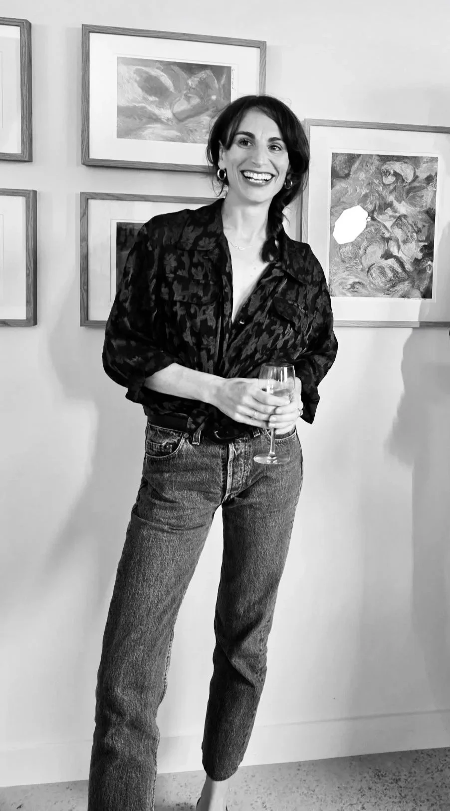 Une femme souriante tenant un verre de vin blanc, portant un chemisier à motifs et un jean, debout devant un mur décoré d'œuvres d'art plastiques.